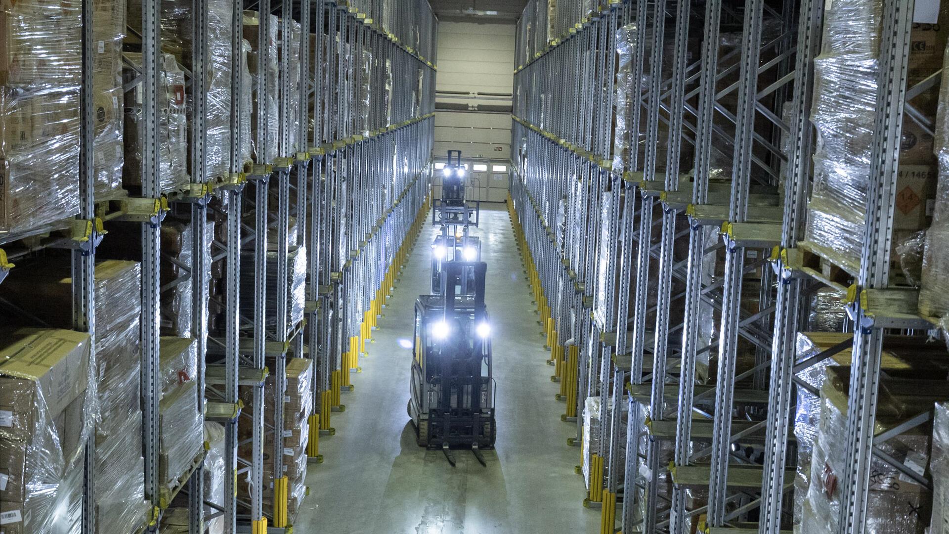 Ein großes, modernes Lager mit hohen Regalen, die bis zur Decke reichen, gefüllt mit verpackten Waren auf Paletten. Mehrere Gabelstapler mit eingeschaltetem Licht fahren durch einen breiten Gang in der Mitte des Lagers.