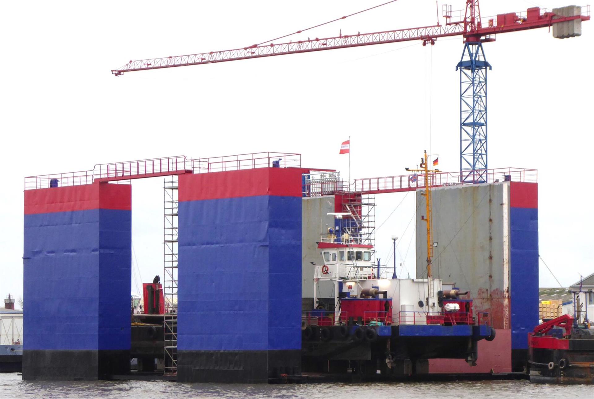 Ein schwimmendes Trockendock mit blau-roter Bemalung im Wasser, vor einem Kran im Hintergrund.