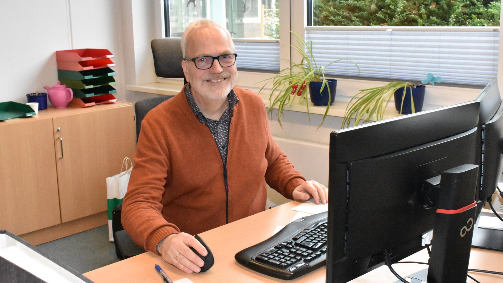 Ein älterer Mann mit einer brauen Jacke sitzt an einem Schreibtisch, auf dem ein Computer steht.