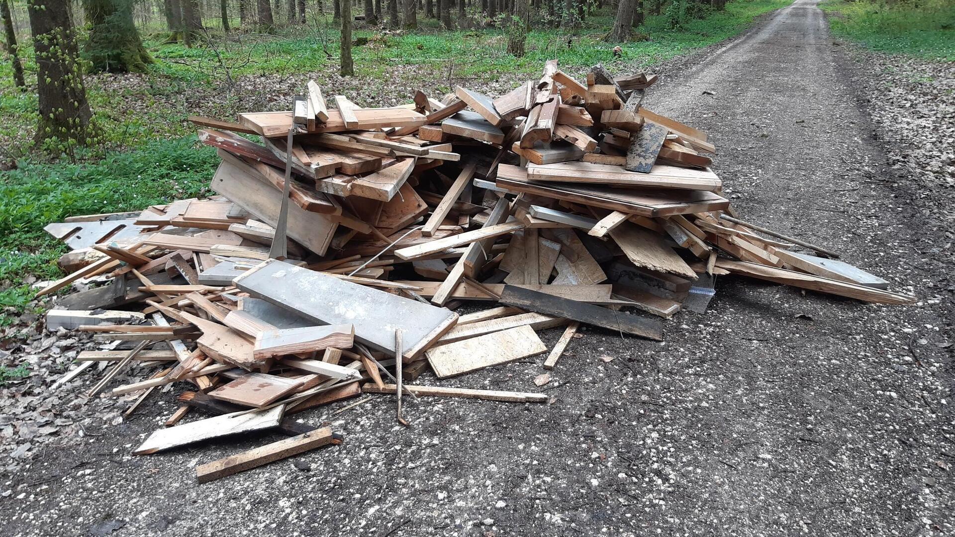 Ein großer Haufen aus Holzabfällen, darunter Bretter, Balken und andere Holzstücke, liegt am Rand eines unbefestigten Waldwegs. Im Hintergrund ist dichter Wald mit grünem Unterwuchs und hohen Bäumen zu sehen.