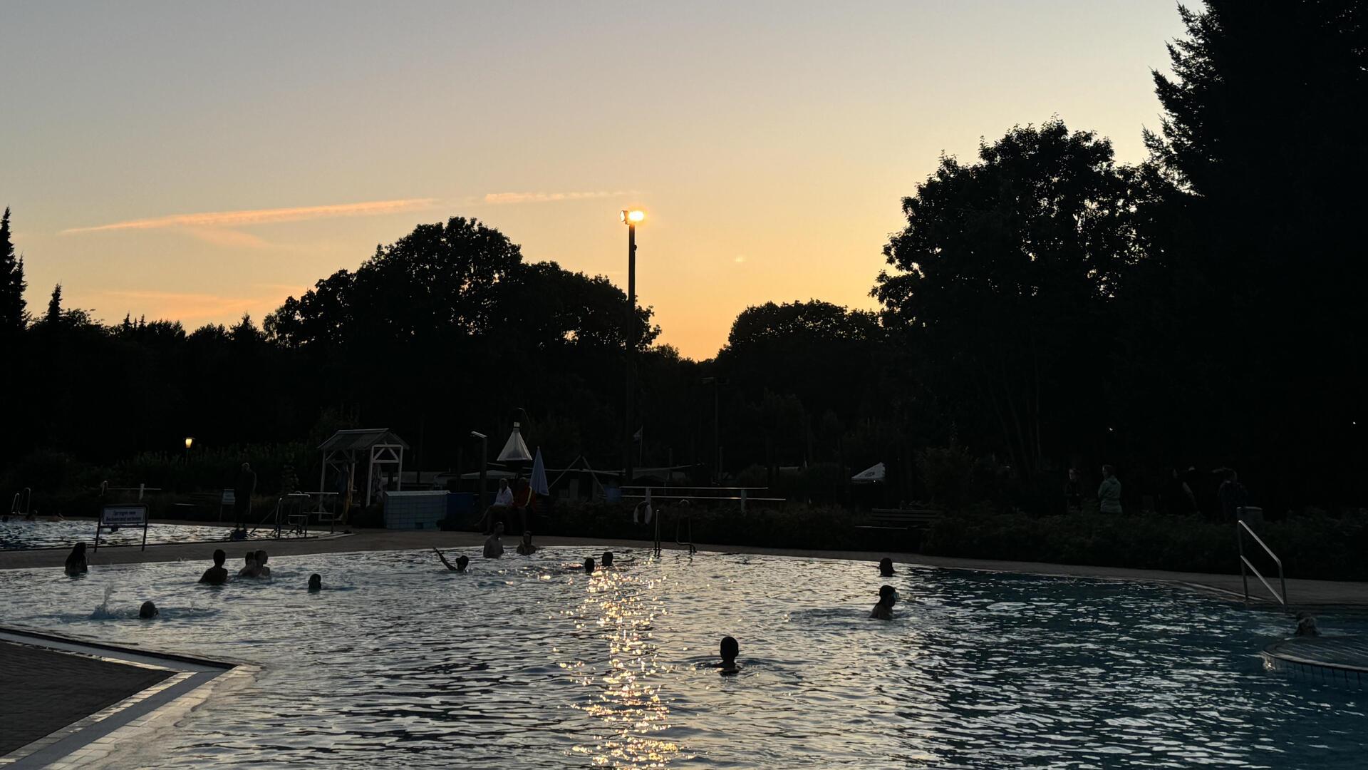 Badegäste schwimmen in einem Freibad, während bereits die Sonne untergeht.