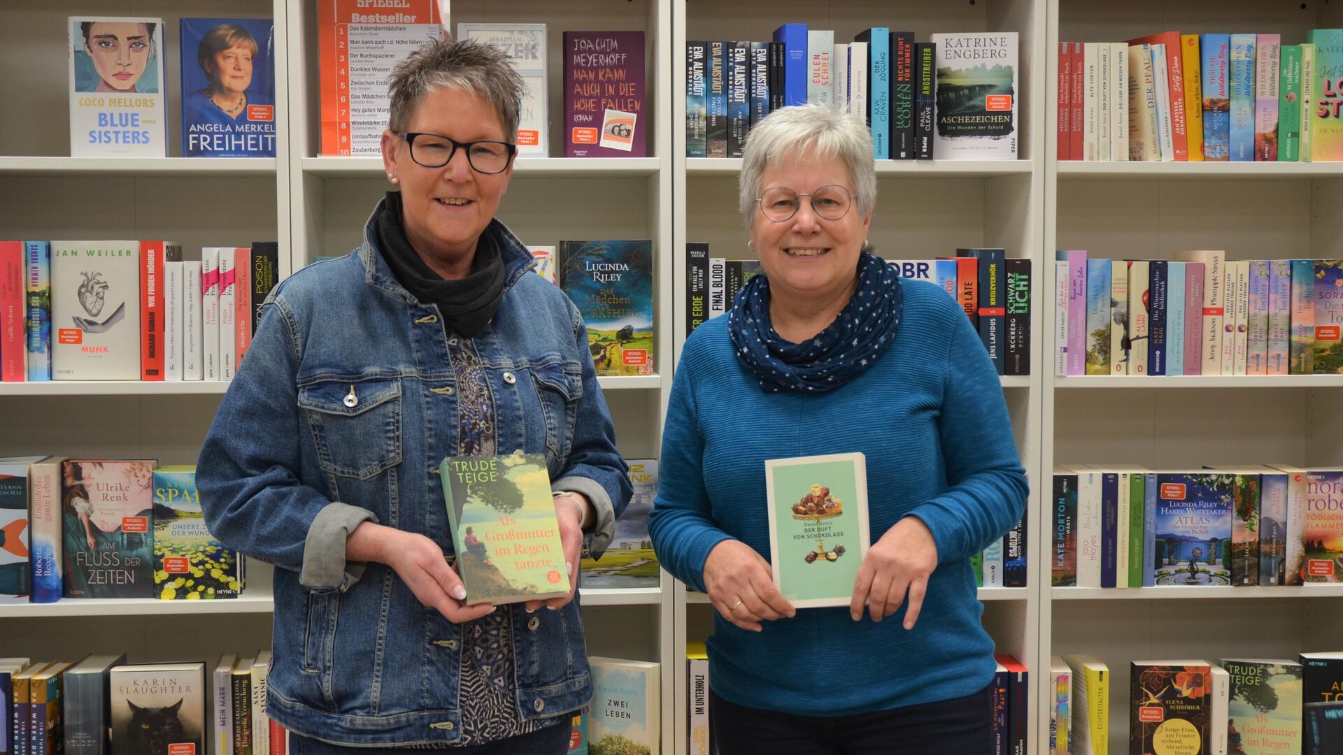 Zwei Frauen mit jeweils einem Buch in der Hand stehen vor einer Bücherwand und lächeln in die Kamera. 