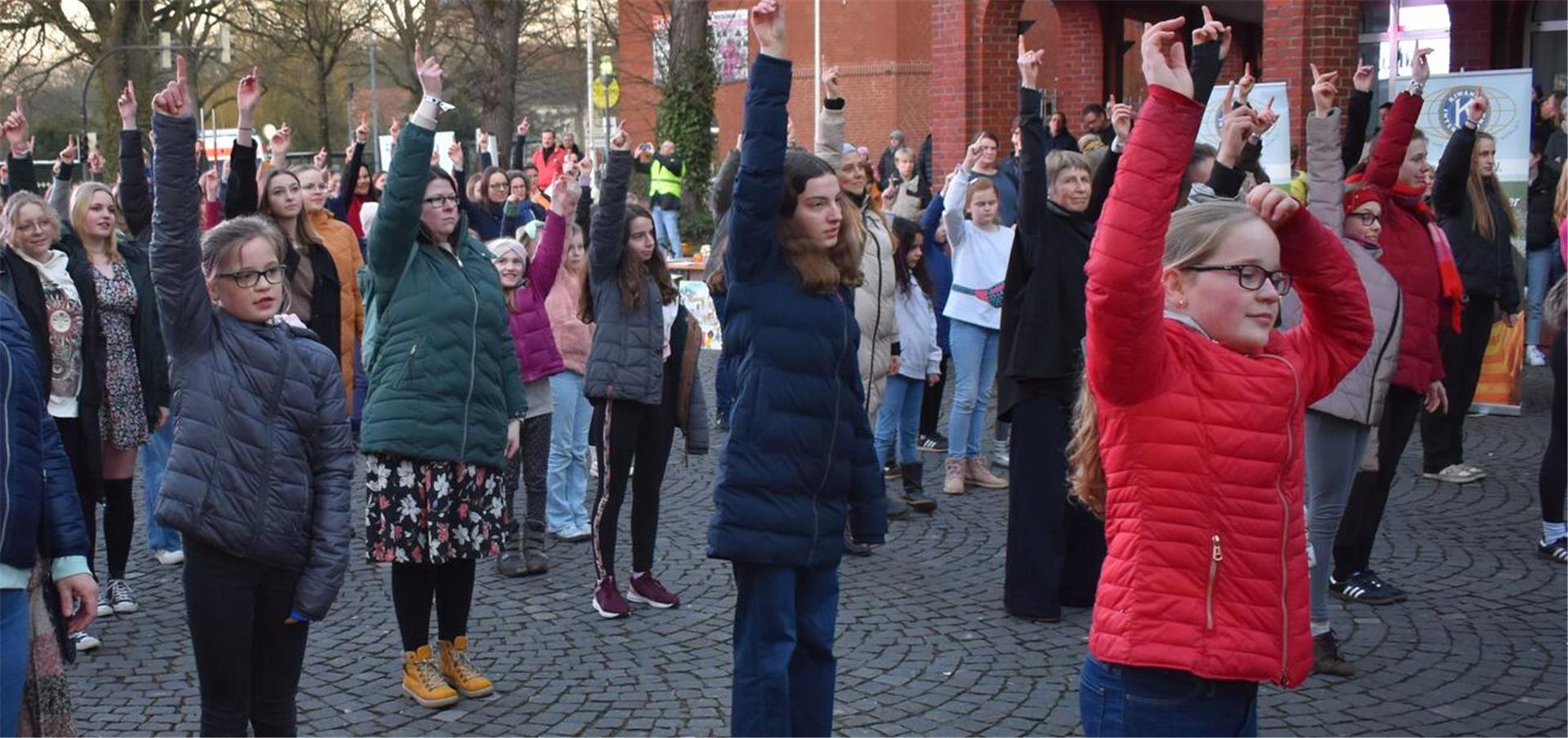 Viele Kinder und Erwachsene stehen auf einem Platz und heben den rechten Arm hoch.