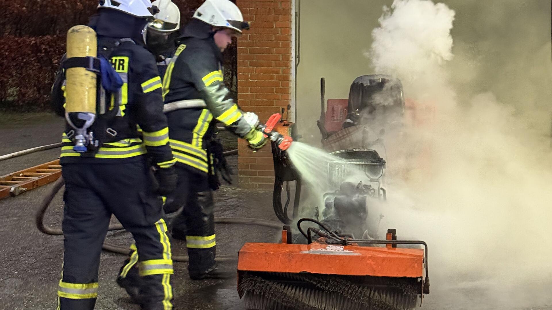 Drei Männer in schwarz- und signalgelben Anzügen halten einen Wasserstrahl auf einen brennenden Aufsitzrasenmäher.