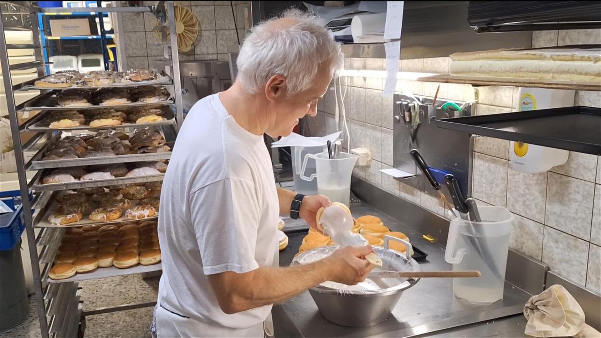 Ein Mann glasiert Berliner in einer Backstube. Im Hintergrund sind viele Kuchen auf Blechen.