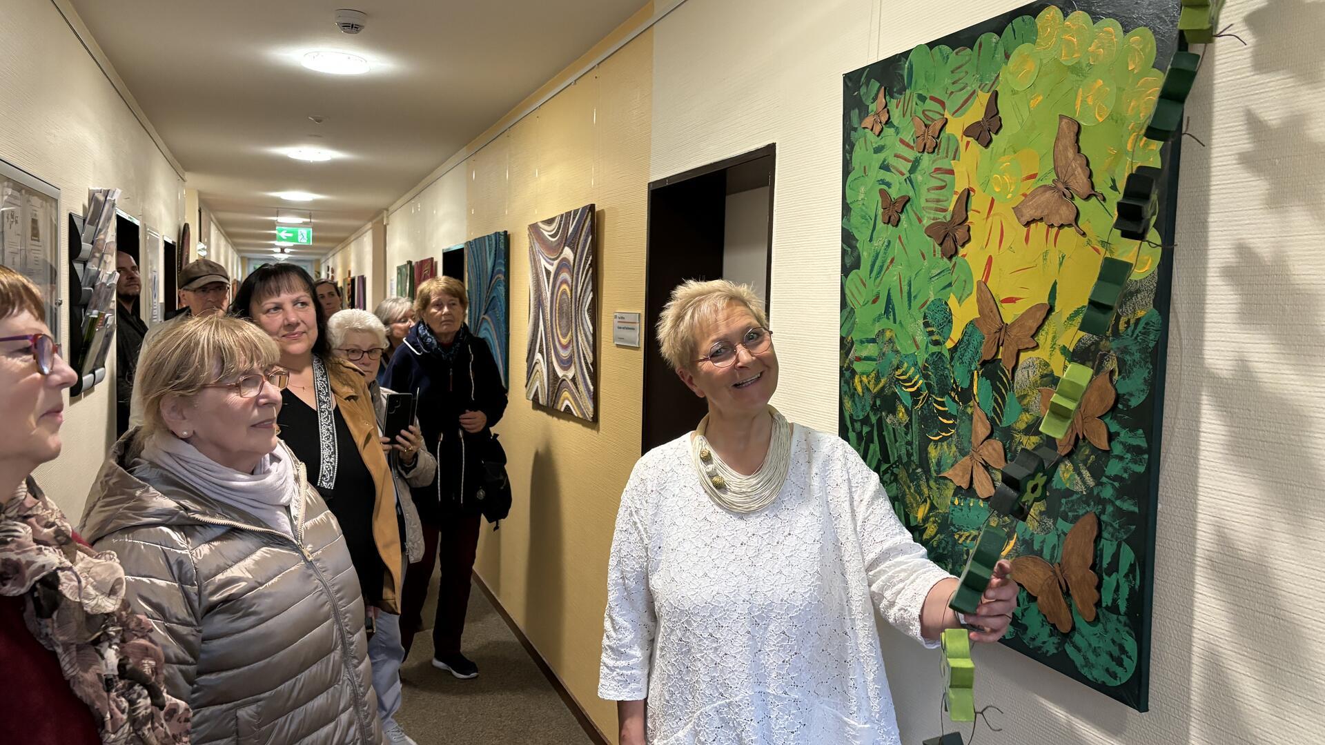 Eine Gruppe von Menschen steht in einem Flur und betrachtet ein Kunstwerk an der Wand. Die Künstlerin, in weiß gekleidet, lächelt und zeigt auf das Bild, das aus grünen und braunen Elementen besteht und Naturmotive darstellt.