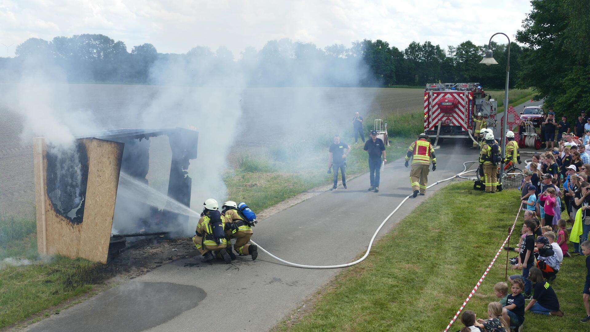 Zwei kniende Feuerwehrleute richten Wasser aus einem Löschschlauch auf einen nur noch rauchenden Holzverschlag. In sicherer Entfernung beobachtet Publikum die Löschung dieses simulierten Zimmerbrands.