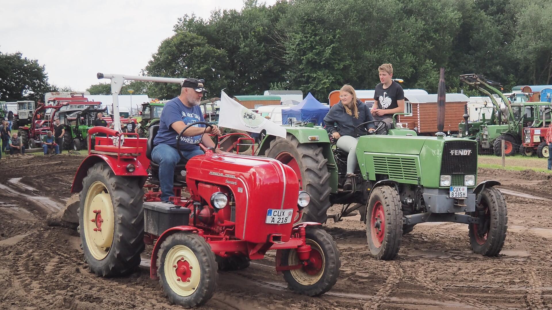 Ein roter Traktor der Marke Porsche und ein grüner Traktor der Marke Fendt werden auf einer Ackerfläche nebeneinander gefahren. 