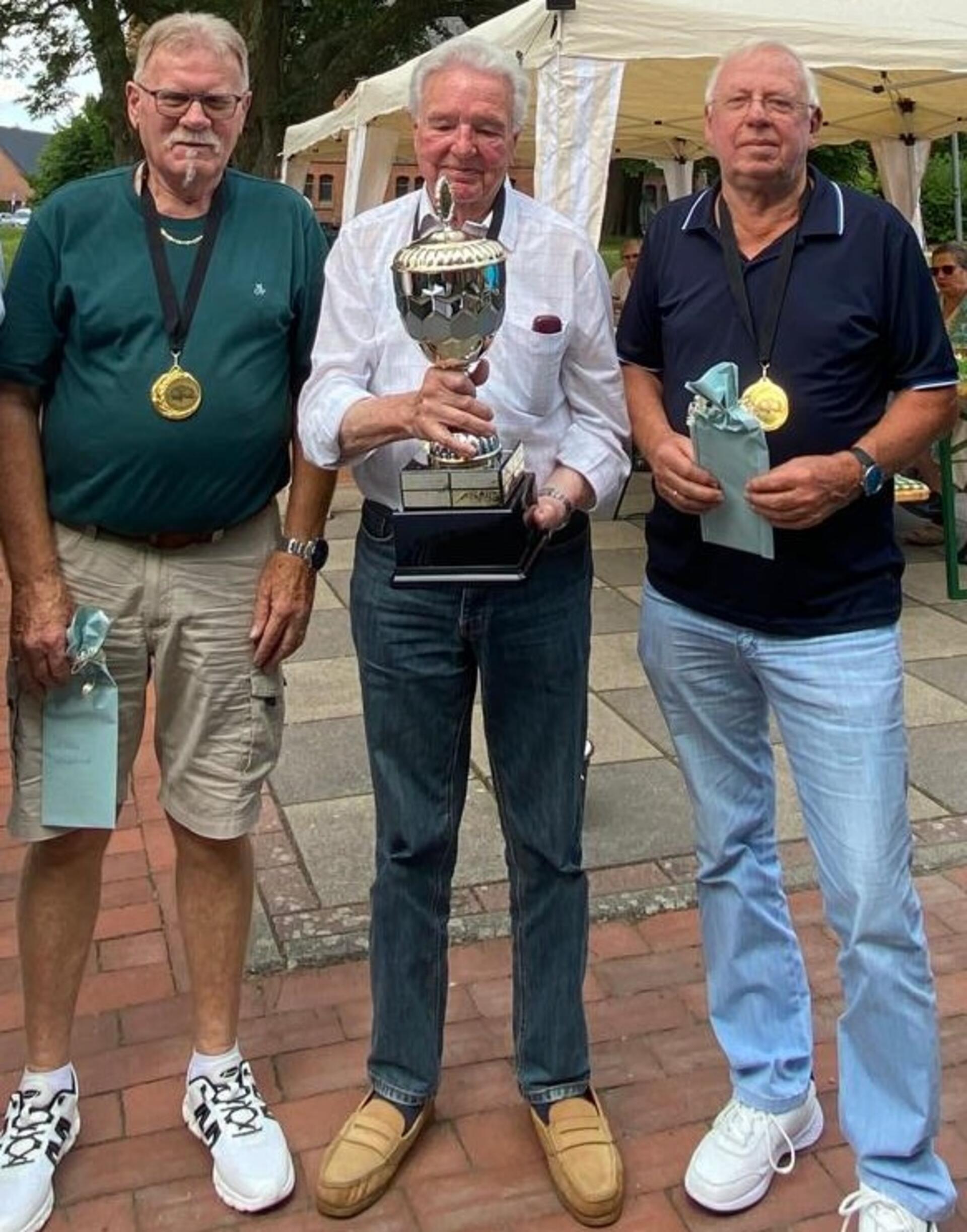 Drei ältere Männer schauen in die Kamera. Der Mann in der Mitte hält einen Pokal in den Händen.