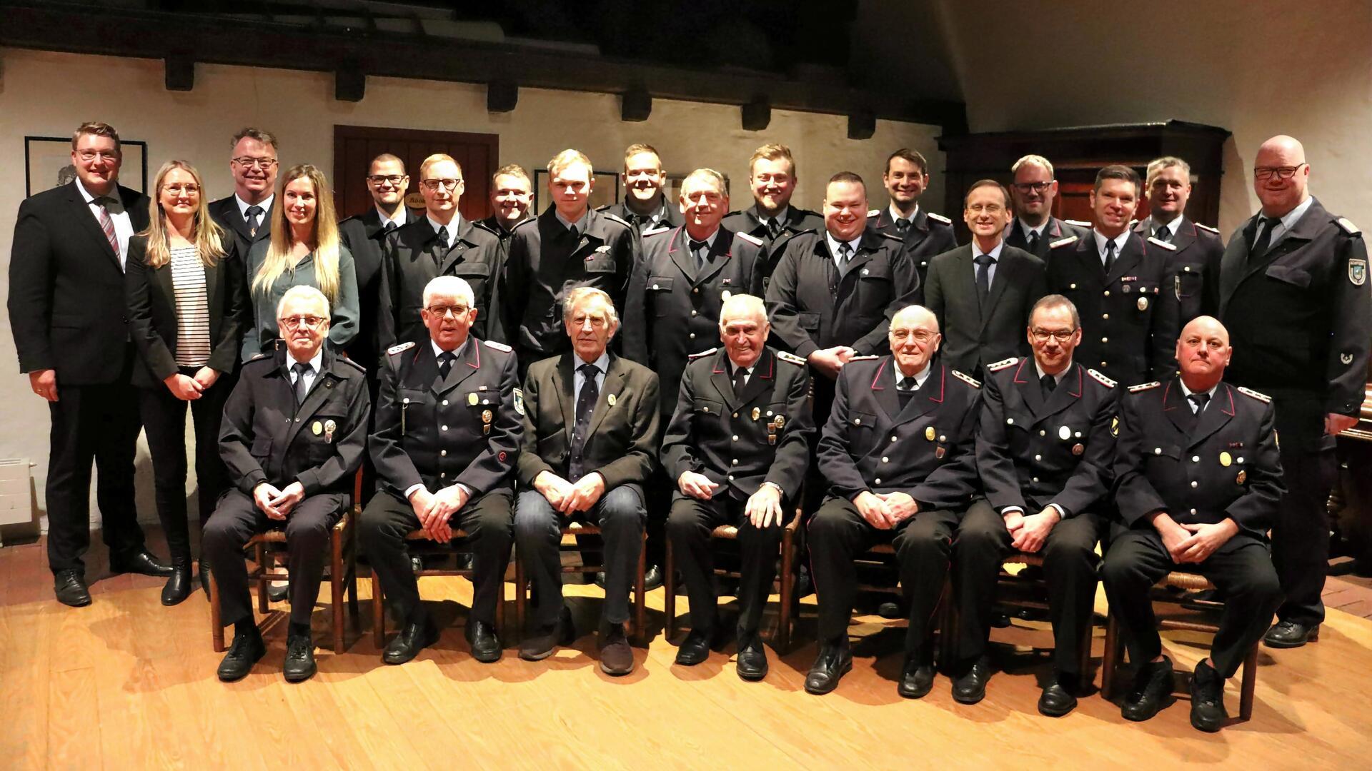 Eine größere Gruppe von Personen posiert für ein Gruppenfoto in einem Innenraum. In der vorderen Reihe sitzen ältere Herren in Feuerwehruniformen, einige mit Auszeichnungen auf der Brust. Hinter ihnen stehen weitere Männer und Frauen, teils in Uniform, teils in formeller Kleidung. 