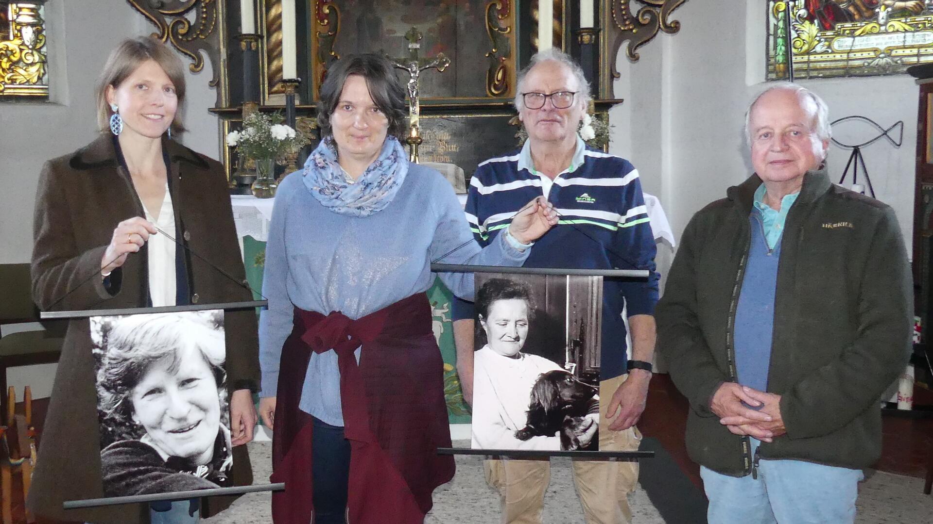 Zwei Frauen und zwei Männer stehen vor dem Altar einer Kirche. Sie halten zwei Schwarz-Weiß-Aufnahmen in Händen, die Porträtfotos zweier Frauen zeigen.