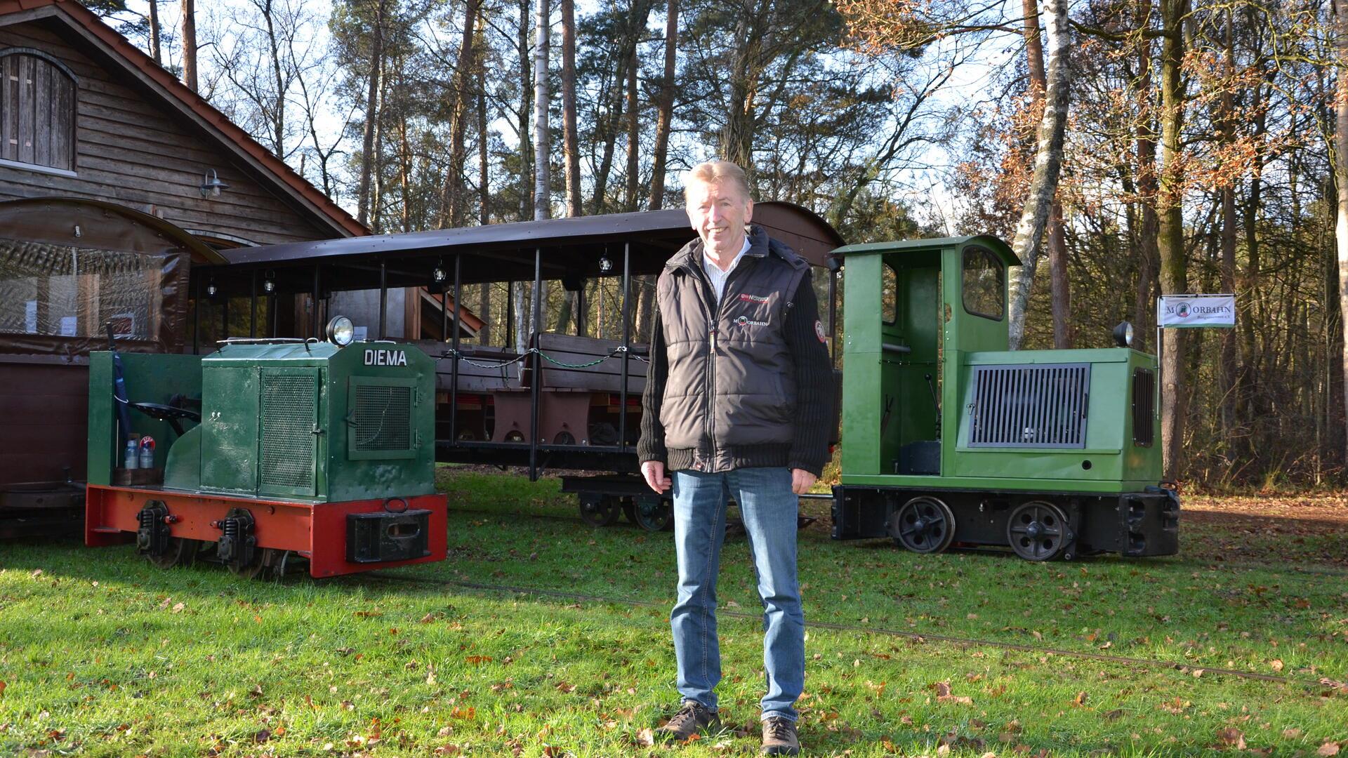 Der Lokführer der Moorbahn steht vor den beiden Loks der Moorbahn am Lokschuppen.