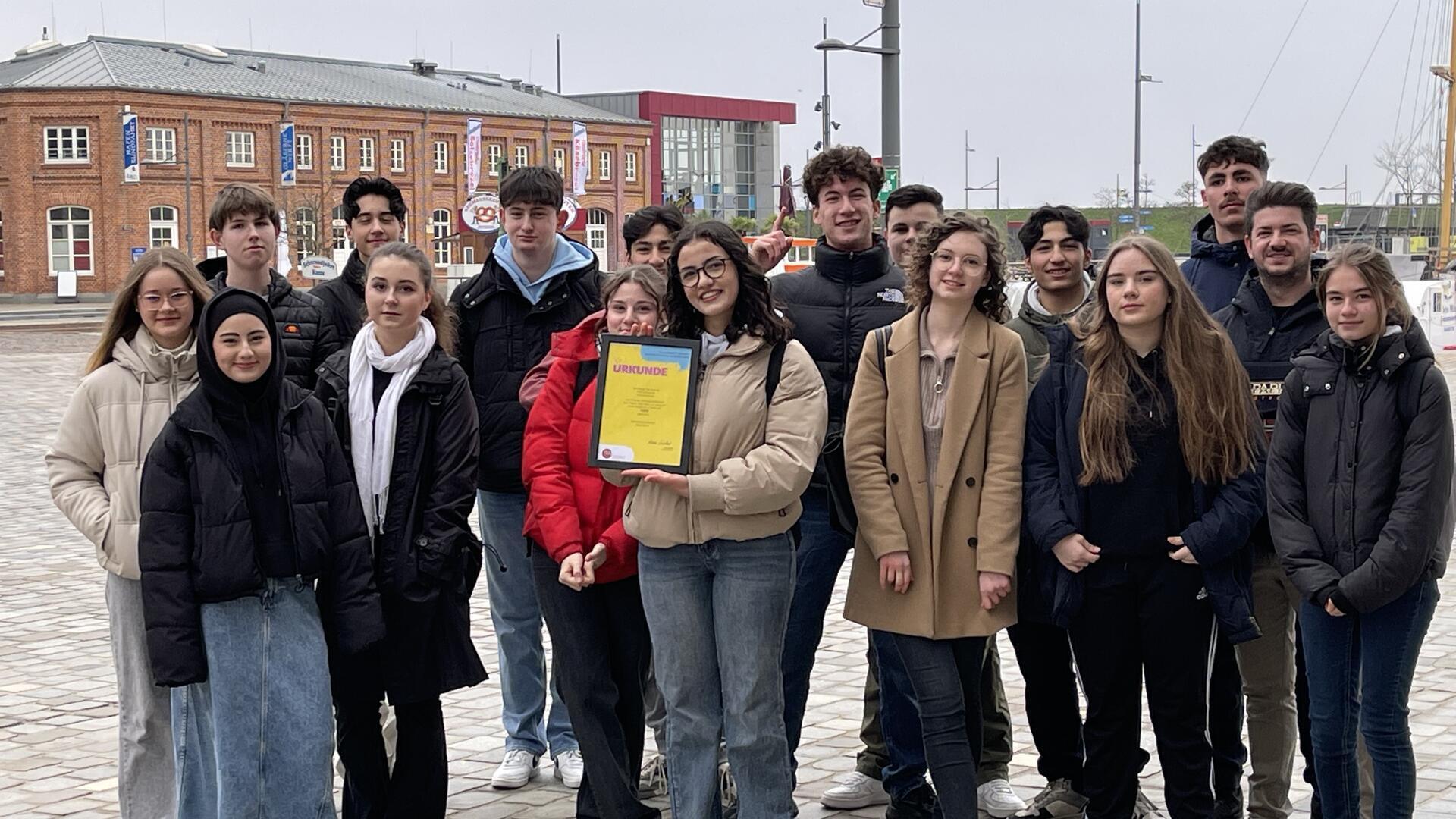 8. Klasse steht zusammen am Neuen Hafen in Bremerhaven, eine Schülerin hält eine gerahmte Urkunde in Händen