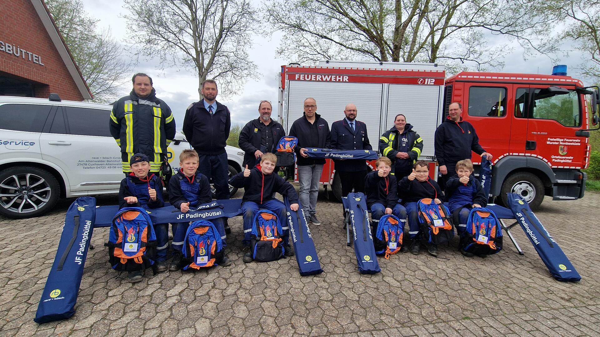 Vor einem Pkw und einem Einsatzfahrzeug der Feuerwehr Padingbüttel posieren sieben Erwachsene und sechs Kinder für ein Gruppenfoto mit neuen Liegen und Rucksäcken.