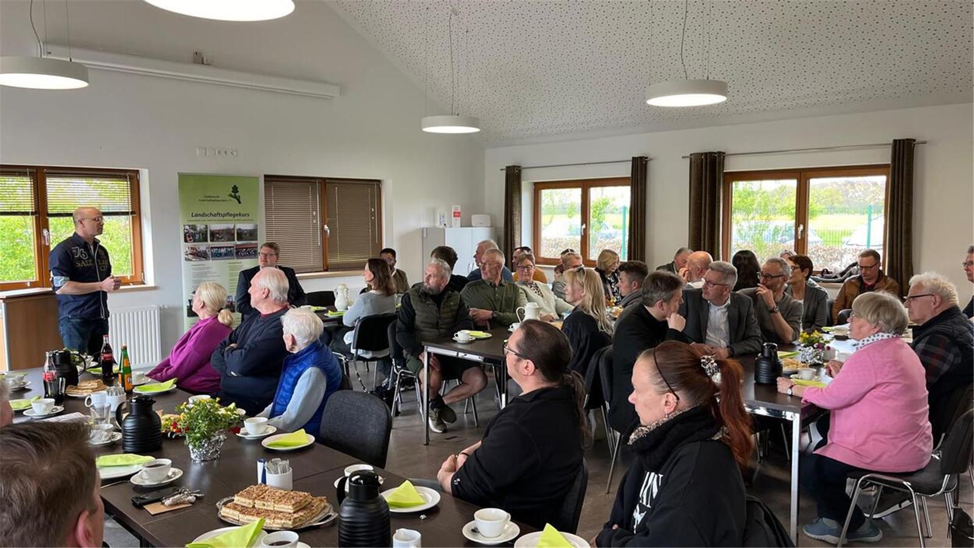 mehrere Männer und Frauen sitzen an langen gedeckten Holztischen, auf denen Kaffee und Butterkuchen zum Verzehr bereitstehen