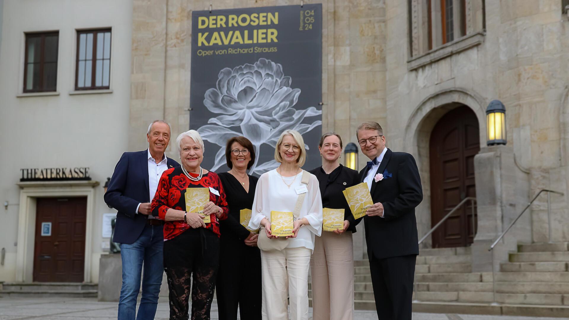 vier Frauen und zwei Männer stehen vor dem Eingang des Stadttheaters Bremerhaven, an dessen Fassade ein großes Plakat hängt mit der Aufschrift "Der Rosenkavalier – Oper von Richard Strauss"