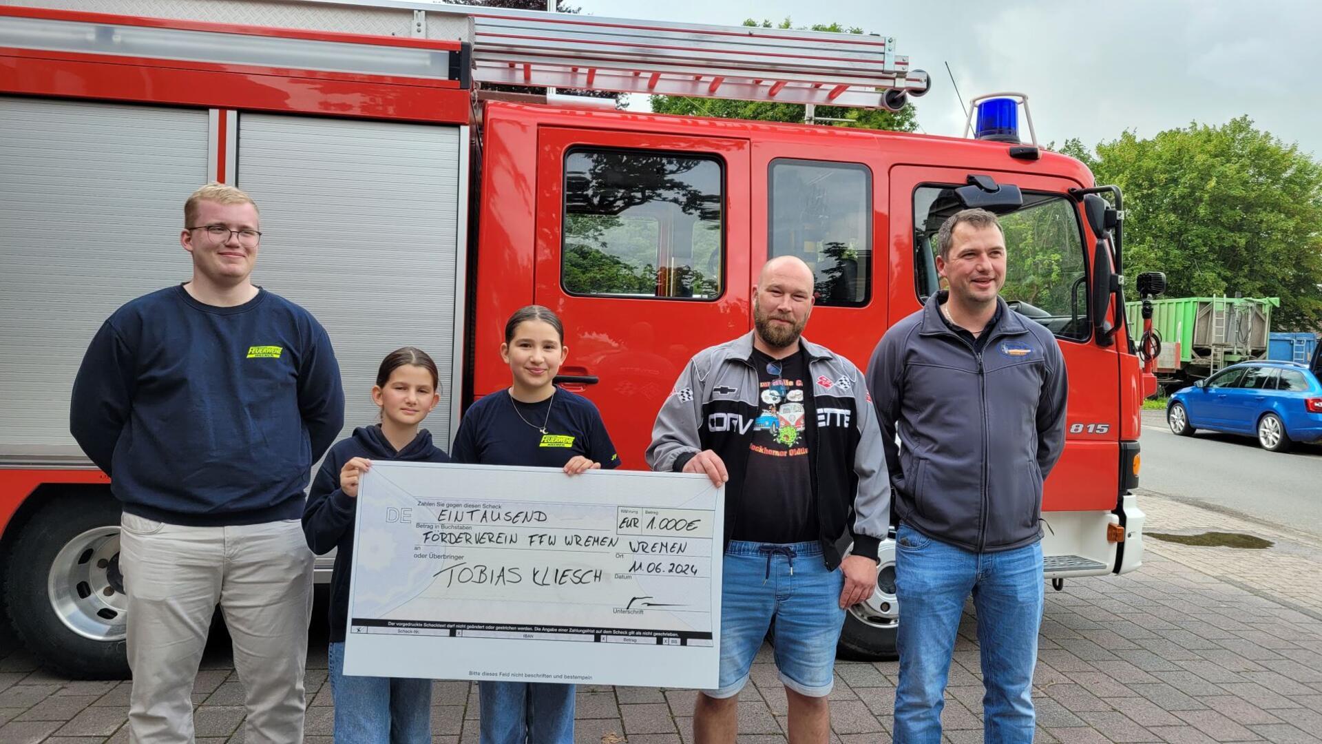 Zwei Kinder und drei Männer stehen nebeneinander vor einem Feuerwehrfahrzeug und halten einen symbolischen Spendenscheck hoch.