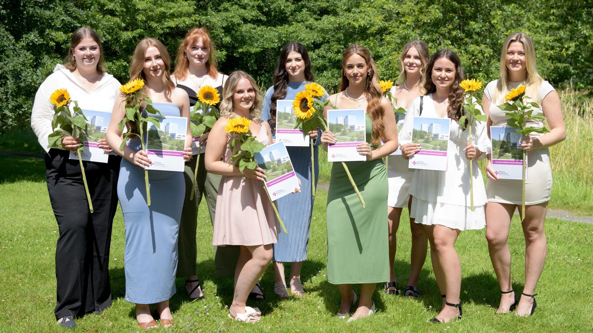 Neun junge Frauen stehen nebeneinander im Grünen. Sie halten ihre Abschlusszeugnisse und Sonnenblumen.