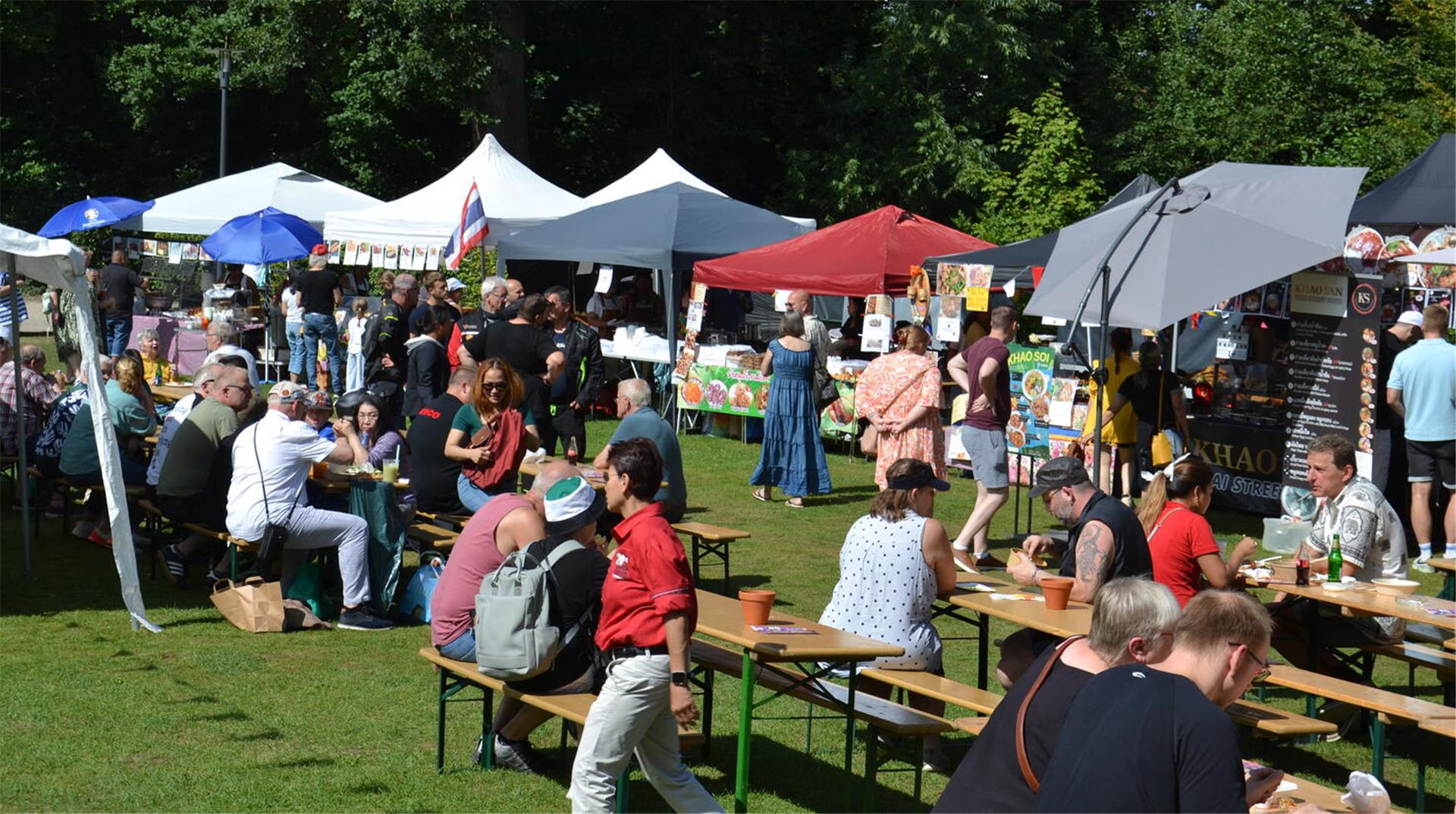 Menschen sitzen an Biertischgarnituren im Zevener Stadtpark. Im Hintergrund Pavillons