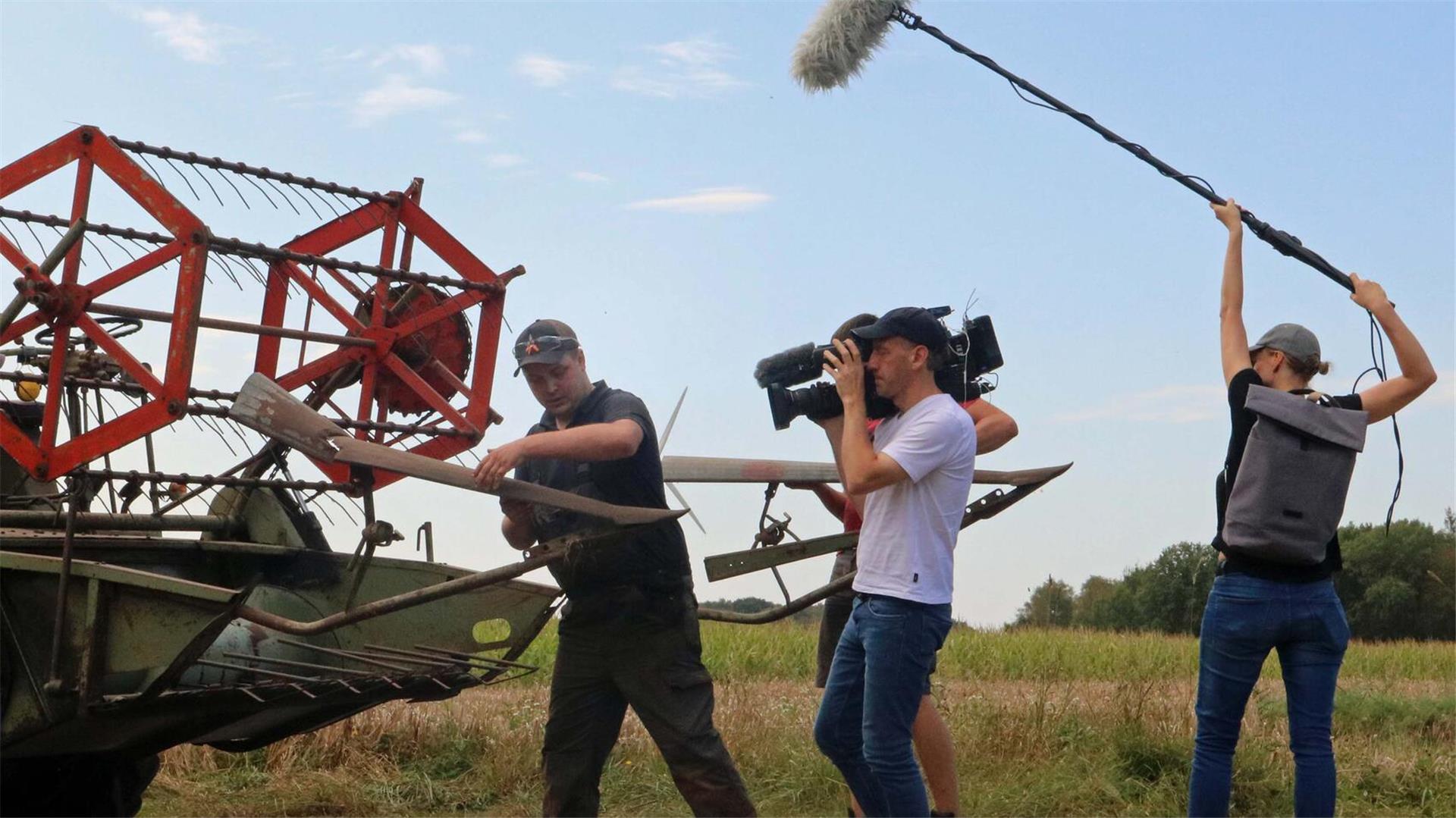 Ein dreiköpfiges Filmteam mit Kamera und Mikrofon filmt einen Mann bei den Arbeiten an einem historischen Mähdrescher auf einem Feld.