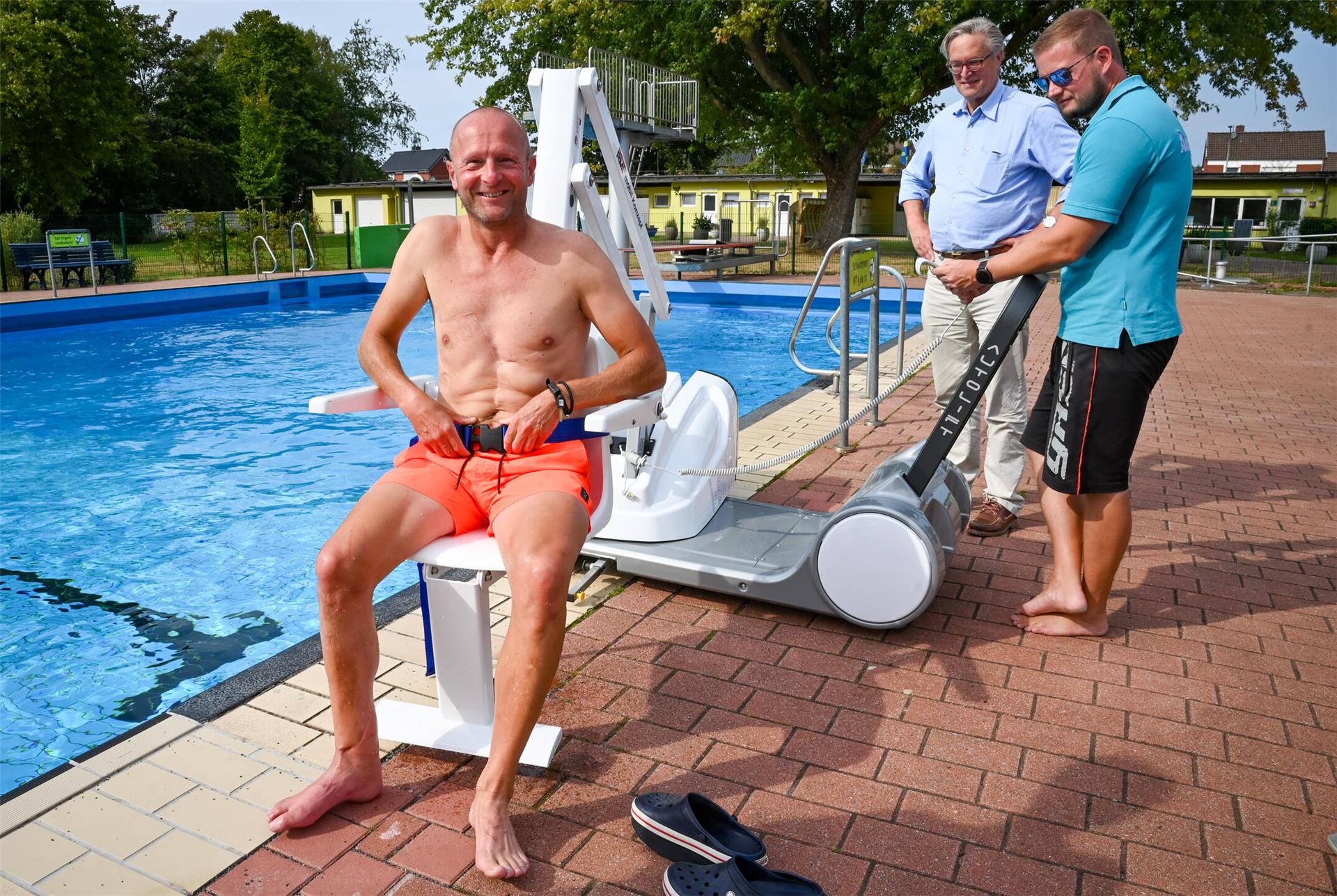 Ein Mann in Badehose nimmt auf einem "Poollifter" am Rande eines Freibad-Beckens Platz. Zwei weitere Männer bedienen das Gerät.