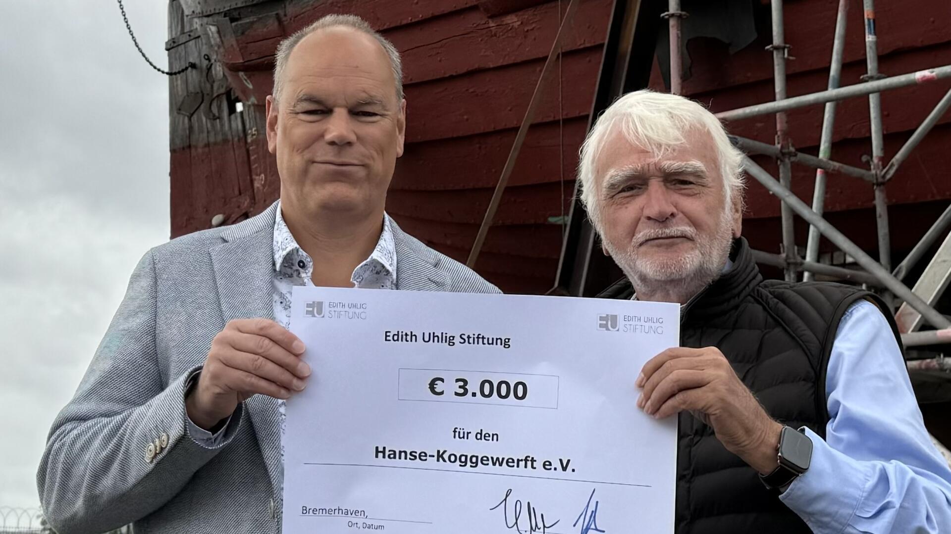 Zwei Männer stehen vor einem eingerüsteten Schiff und halten gemeinsam einen Spendenscheck über 3000 Euro von der Edith-Uhlig-Stiftung an den Verein Hanse-Koggenwerft.