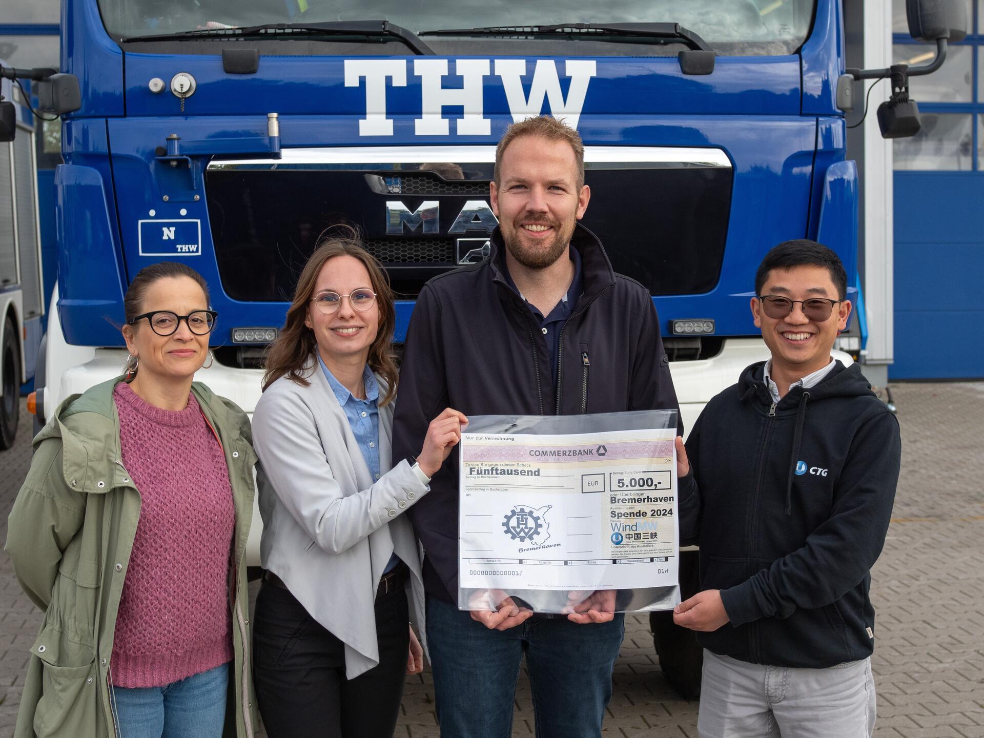 Vier Menschen vor einem blauen THW-Fahrzeug, Spendenscheck in der Hand