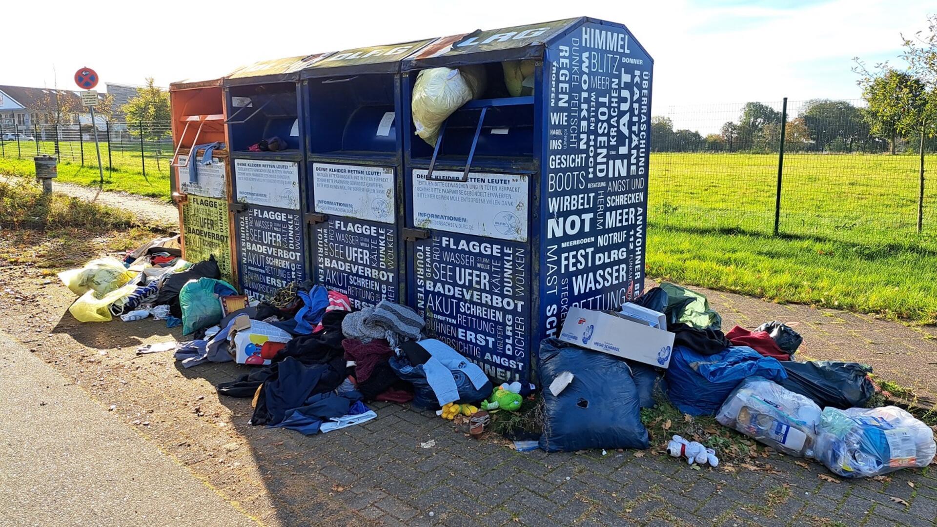 Mehrere Altkleider-Container stehen auf einem Gehweg, umgeben von verstreuten Kleidungsstücken und Plastiktüten sowie Kartons voller Kleidung. 