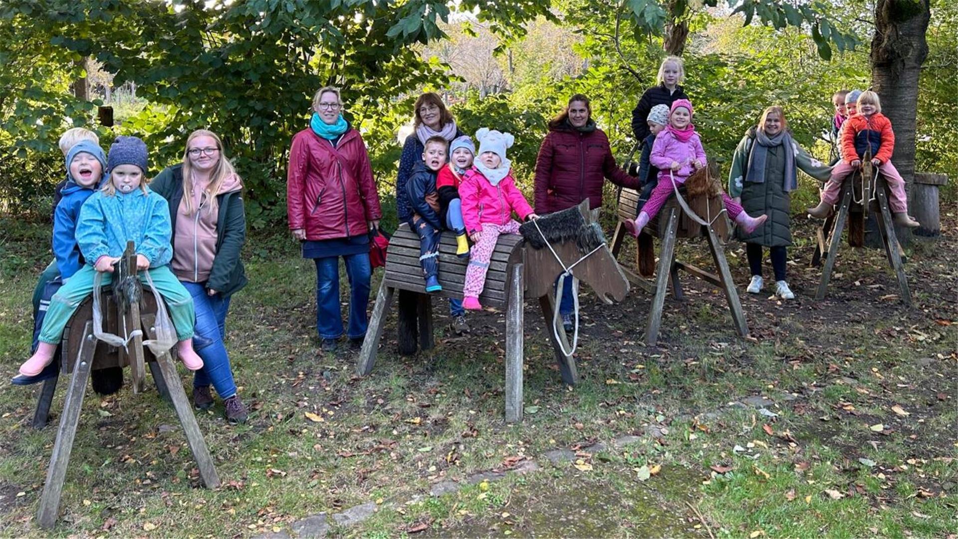 Eine Gruppe von Kindern sitzt auf hölzernen Spielpferden, die in einer Wiese unter Bäumen aufgestellt sind. Jedes Kind trägt warme Kleidung und Mützen, passend für kühleres Wetter. Hinter den Kindern stehen mehrere Erwachsene, die sie begleiten.