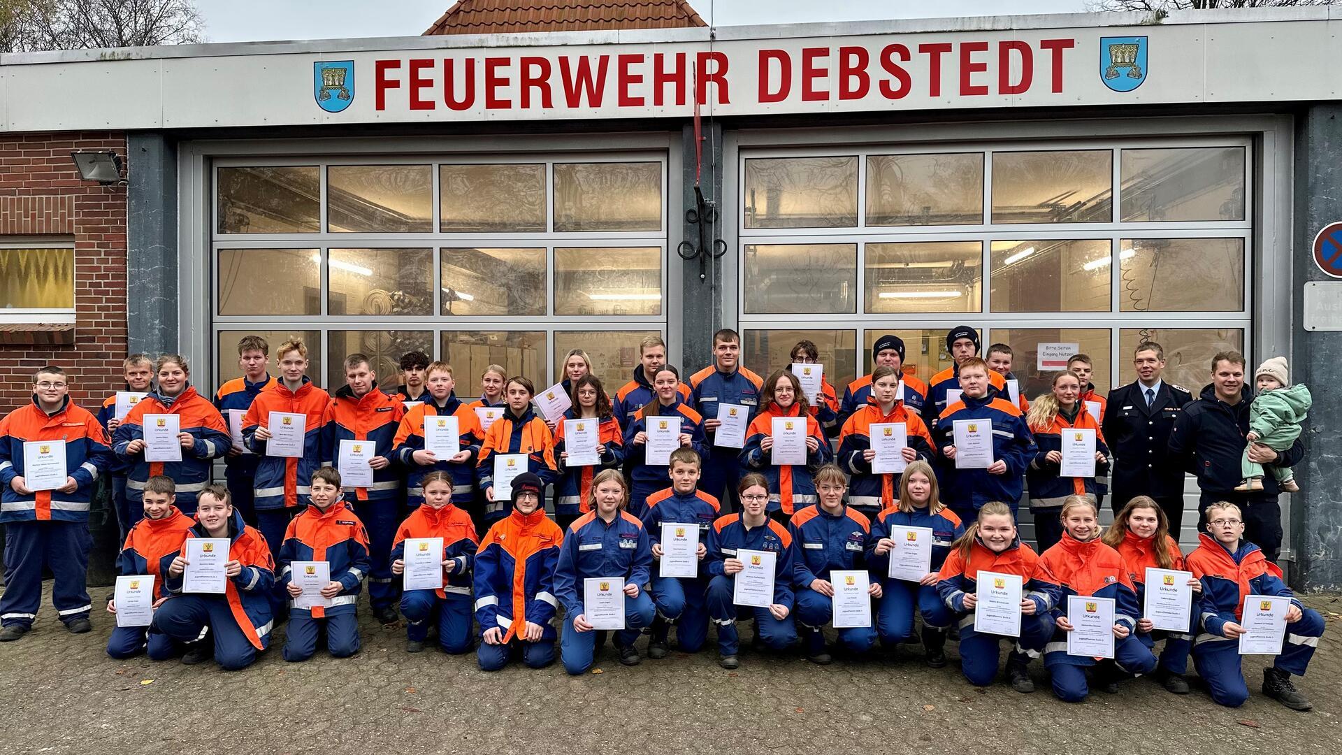 Eine große Gruppe von Kindern und Jugendlichen in orange-blauen Feuerwehruniformen steht vor einem Gebäude mit der Aufschrift „FEUERWEHR DEBSTEDT“. Sie halten stolz Urkunden in den Händen. 