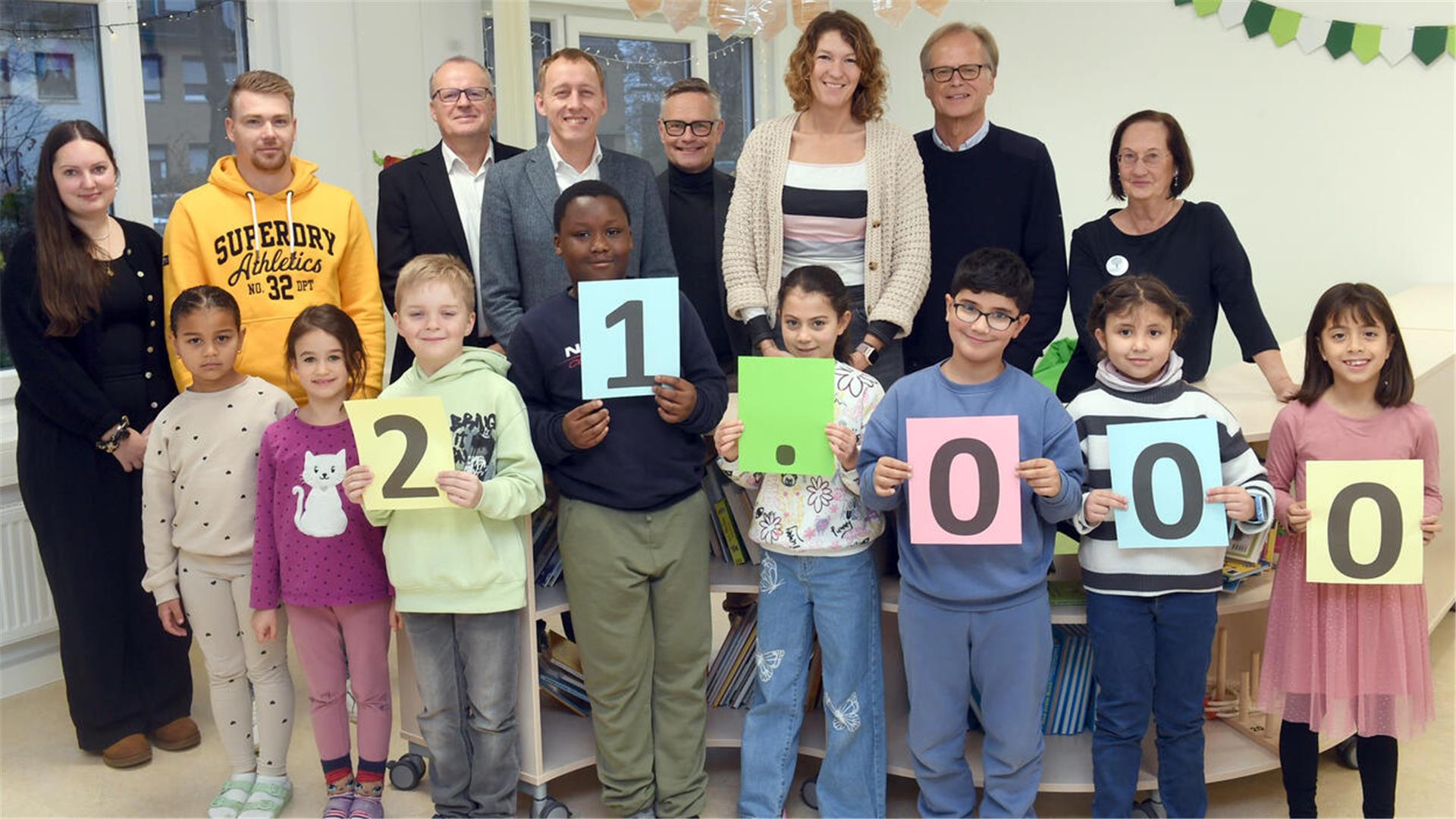 
Eine Gruppe von Kindern und Erwachsenen posiert gemeinsam in einem hellen Raum. Die Kinder stehen in der vorderen Reihe und halten bunte Schilder mit den Ziffern und Zeichen „21.000“. Hinter ihnen stehen die Erwachsenen, die freundlich in die Kamera lächeln. 