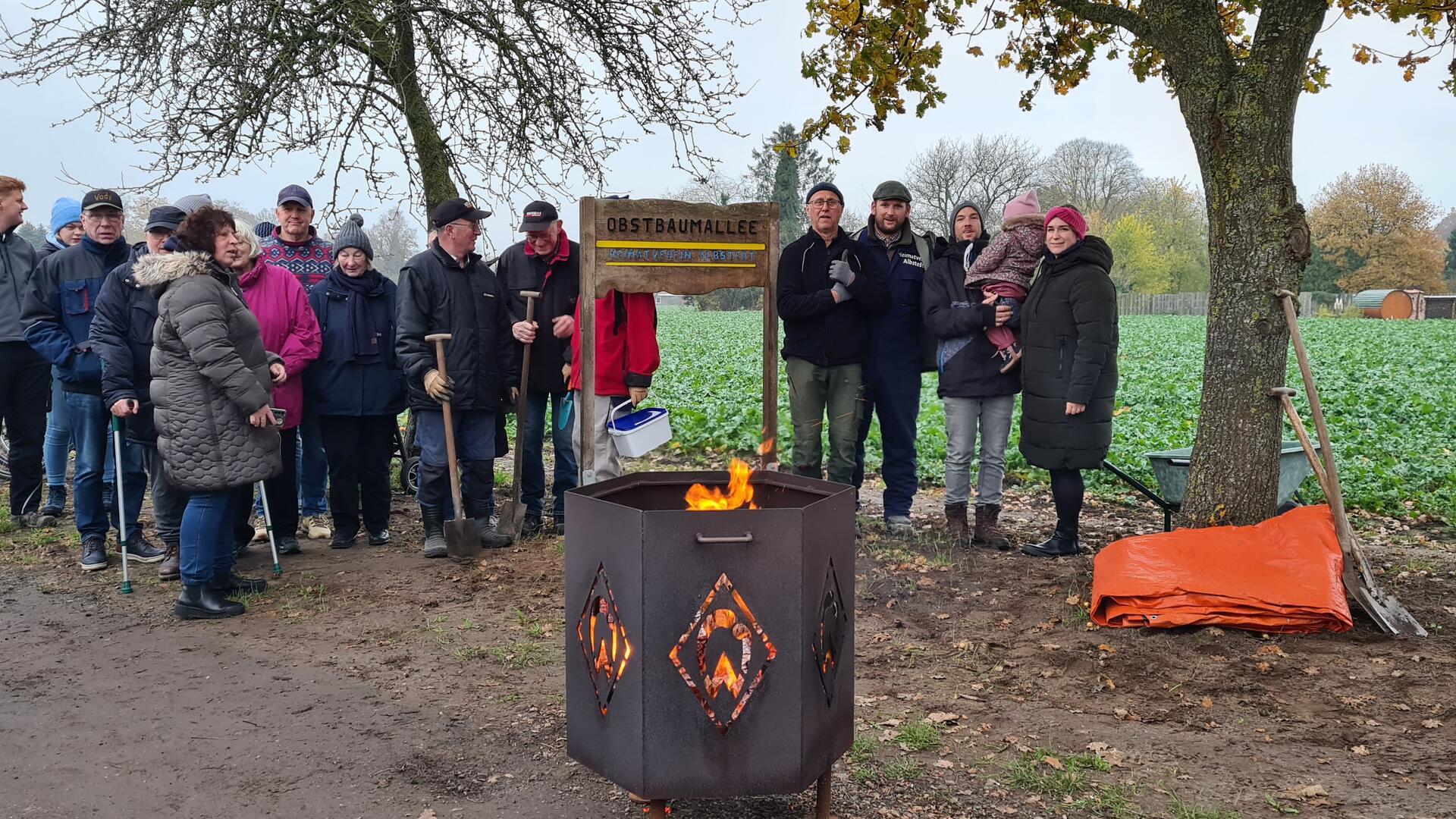 Eine Gruppe von Menschen steht unter Bäumen vor einem Schild mit der Aufschrift „Obstbaumallee“. Im Vordergrund brennt ein Feuer in einer Metalltonne.