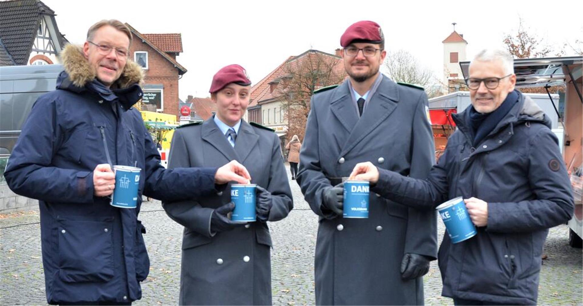 Vier Personen schauen in die Kamera. In der Mitte stehen ein Soldat und eine Soldatin in Uniform. Sie halten eine Spendendose in der Hand. Jeweils daneben steht ein Mann, der Geld in die Spendendose der Soldaten steckt.  