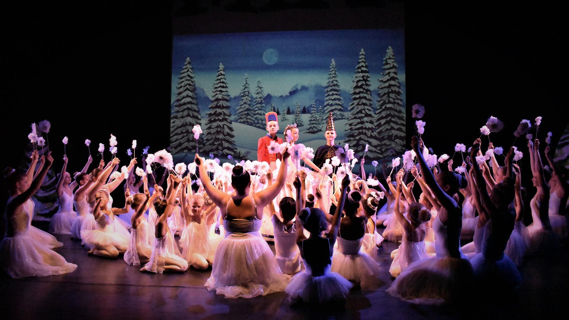 Ballettschüler in weißen Kostümen knien vor einem Tänzer, der als Nussknacker verkleidet ist und halten Blumen in die Höhe.