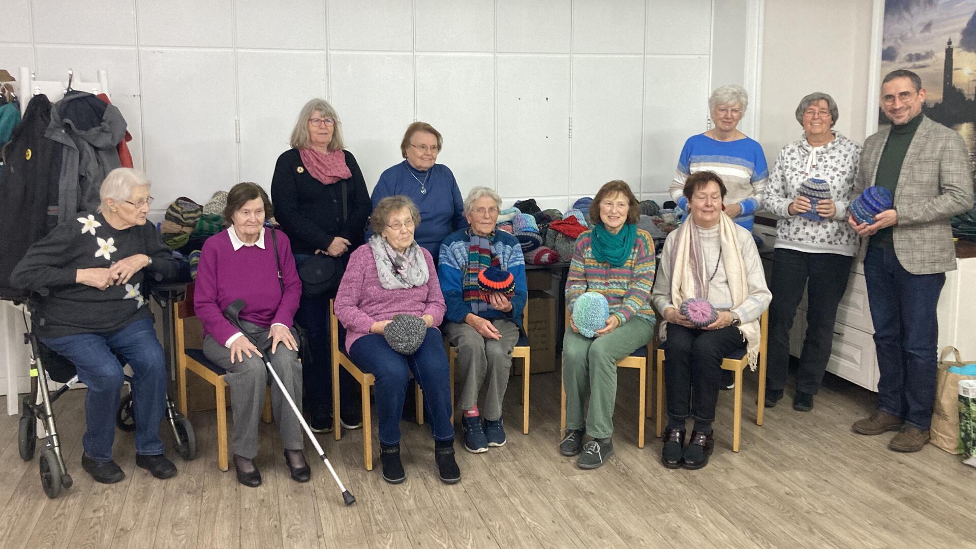 Eine Gruppe älterer Menschen sitzt und steht in einem Gemeinschaftsraum. Einige halten gestrickte Mützen in den Händen. Im Hintergrund sind viele weitere gestrickte Kleidungsstücke auf einem Tisch gestapelt.