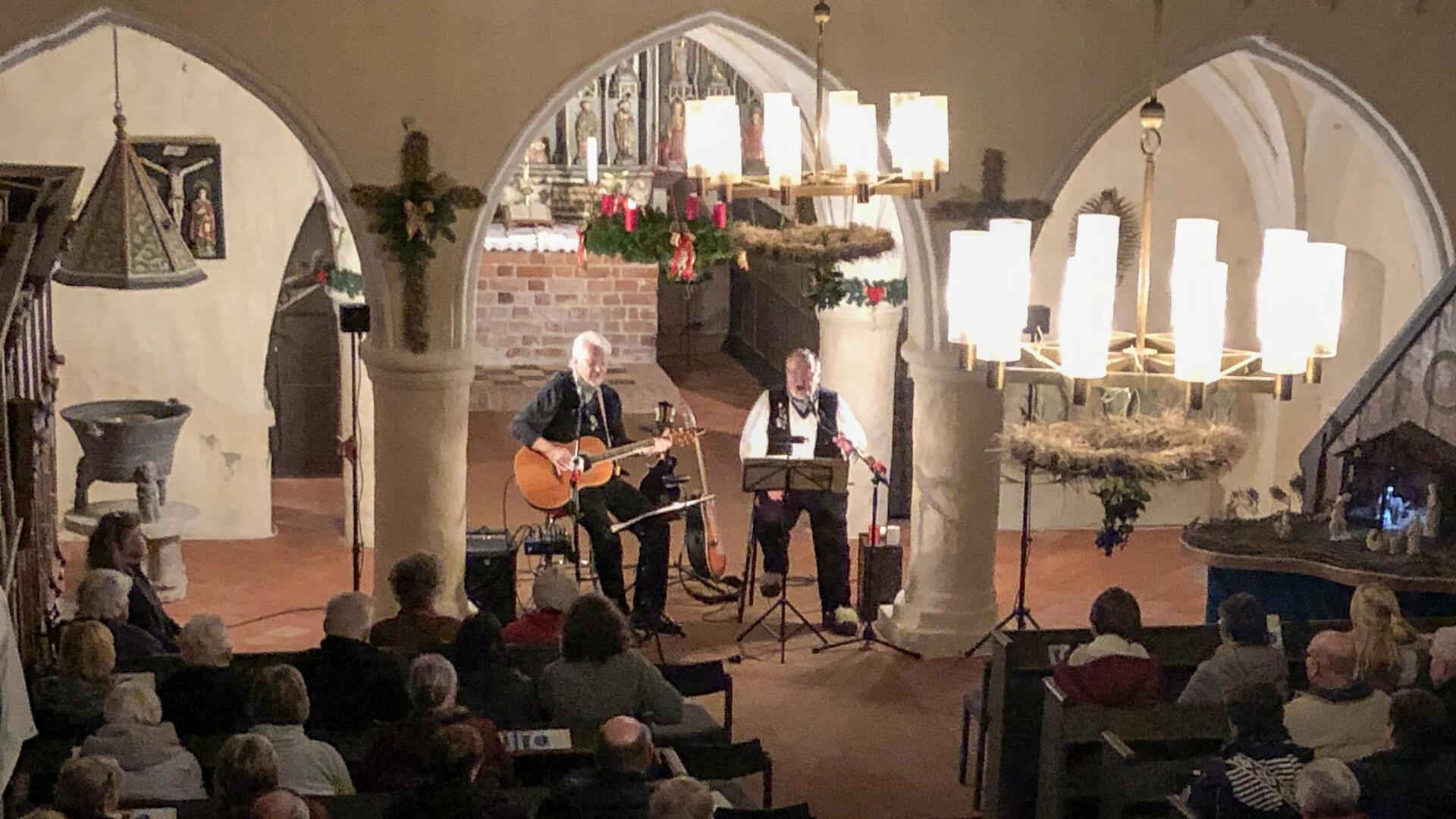 Ein stimmungsvoll beleuchtetes Konzert in einer historischen Kirche. Zwei Musiker spielen, einer mit Gitarre, der andere singend. Im Vordergrund sitzen Zuschauer in Kirchenbänken.