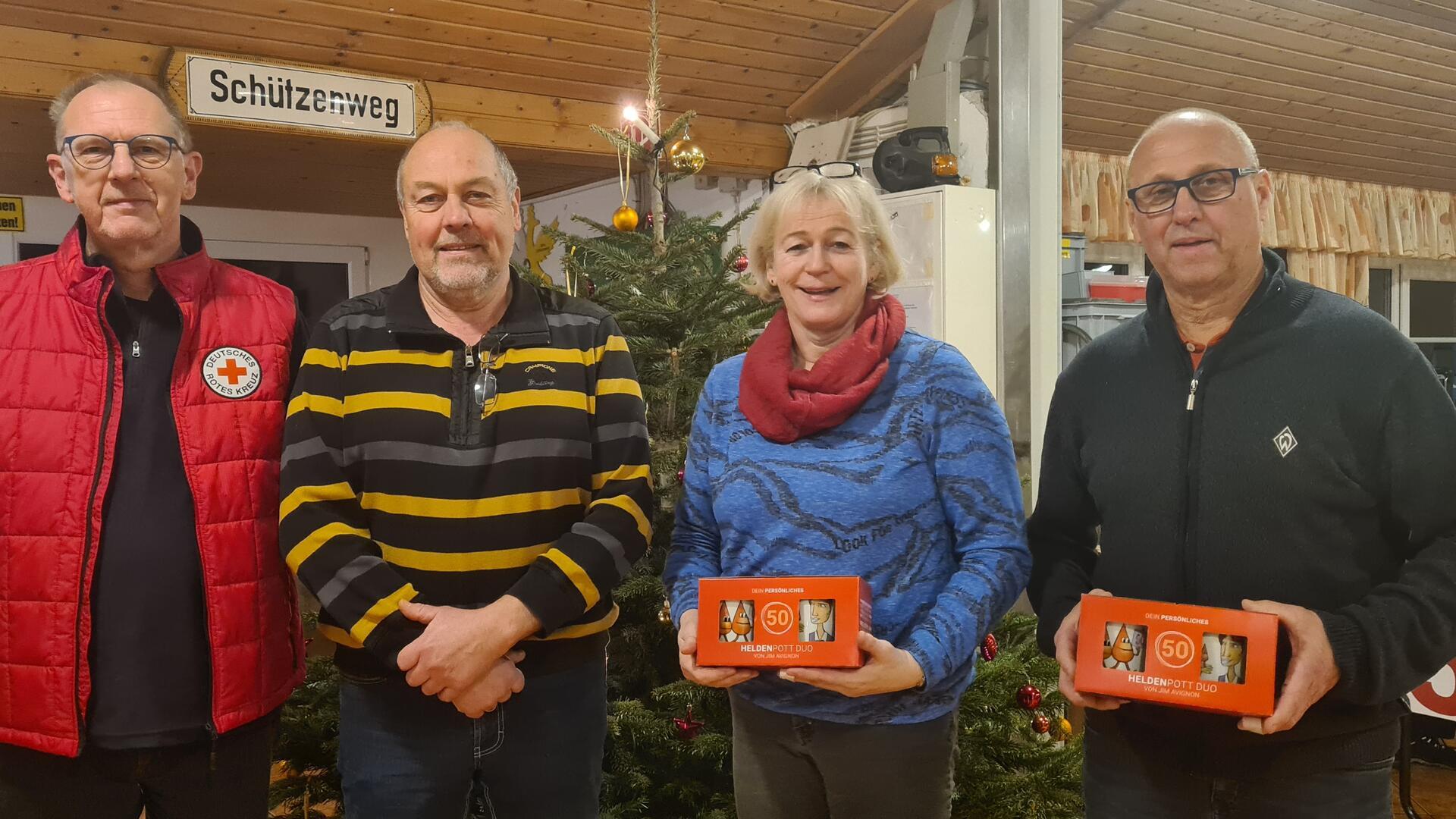 Vier Personen stehen nebeneinander vor einem geschmückten Weihnachtsbaum in einem Raum mit Holzdecke. Links trägt eine Person eine rote Jacke mit dem Logo des Deutschen Roten Kreuzes.