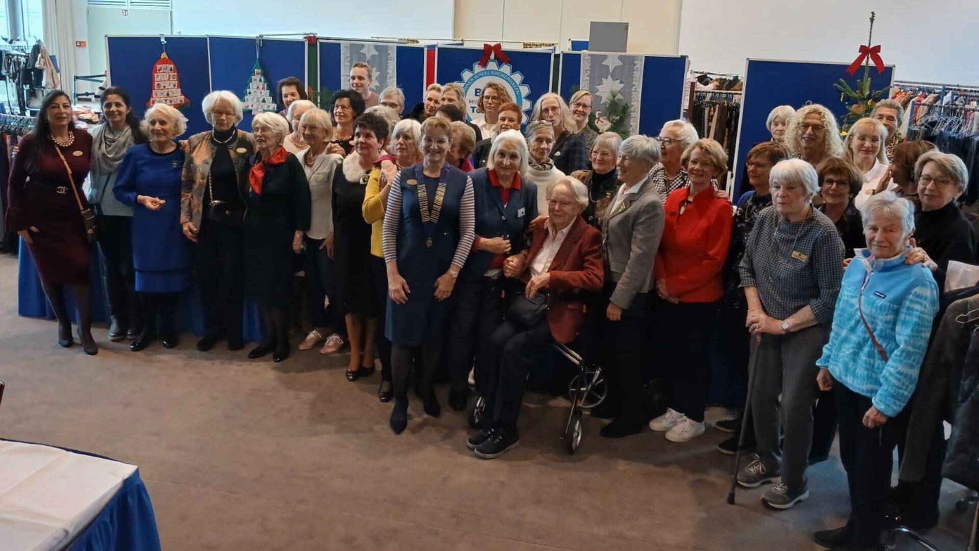 Eine Gruppe von Frauen und einigen Männern unterschiedlichen Alters posiert für ein Gruppenfoto in einem festlich dekorierten Raum. Im Hintergrund sind blau-weiße Stellwände mit weihnachtlichen Verzierungen und Kleidungsauslagen zu sehen.