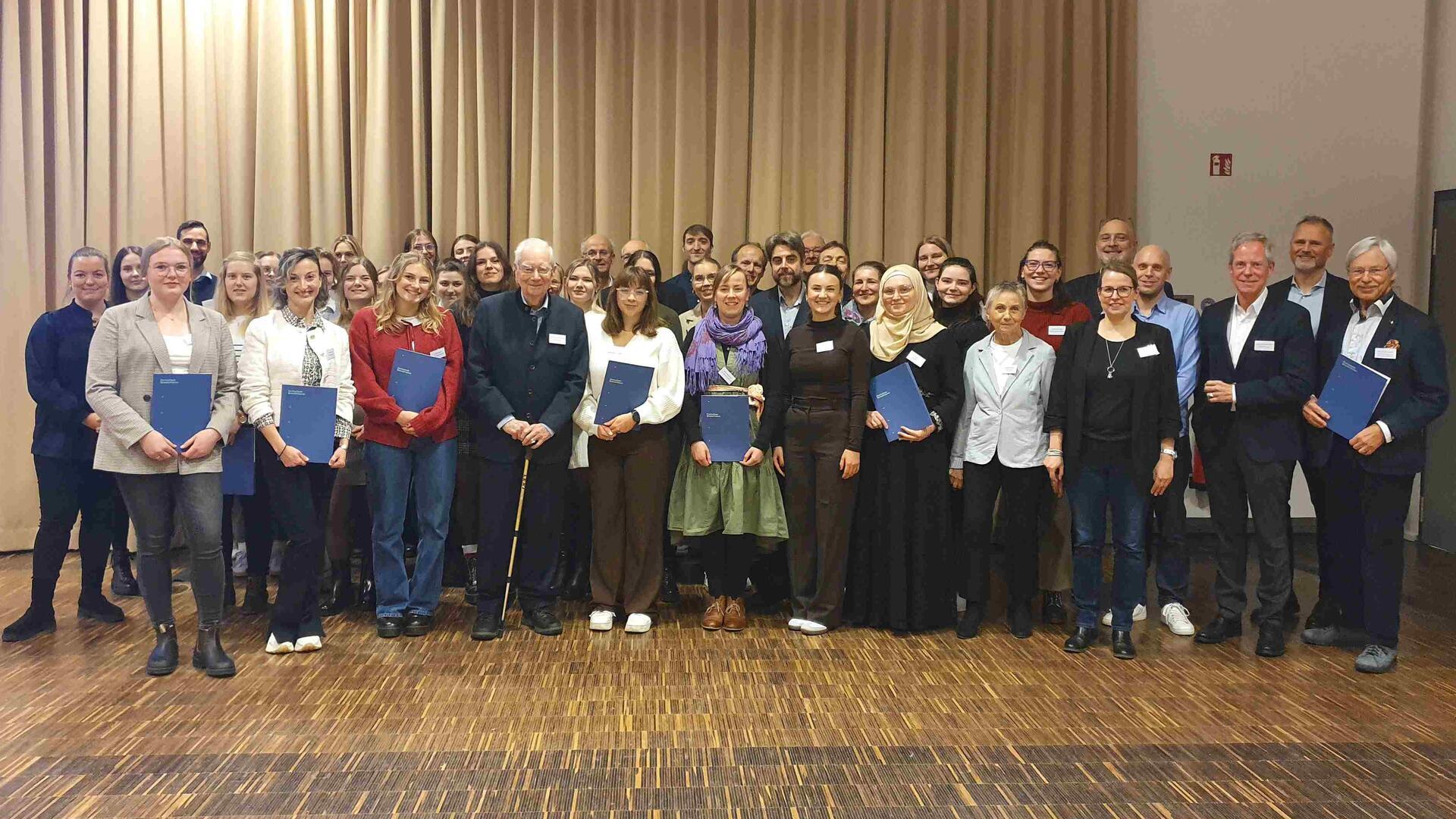 Ein Gruppenfoto einer großen Gruppe von Menschen, die vor einem beigen Vorhang stehen. Viele Personen halten blaue Mappen in den Händen