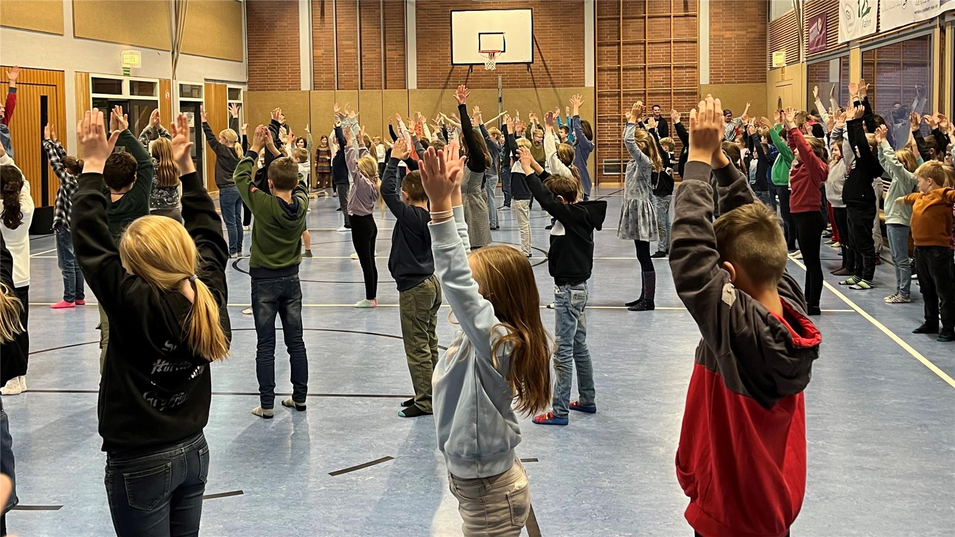 Eine große Gruppe von Kindern und Jugendlichen steht in einer Turnhalle und führt gemeinsam eine Übung aus. Alle haben die Arme nach oben gestreckt, während sie in Reihen aufgestellt sind.