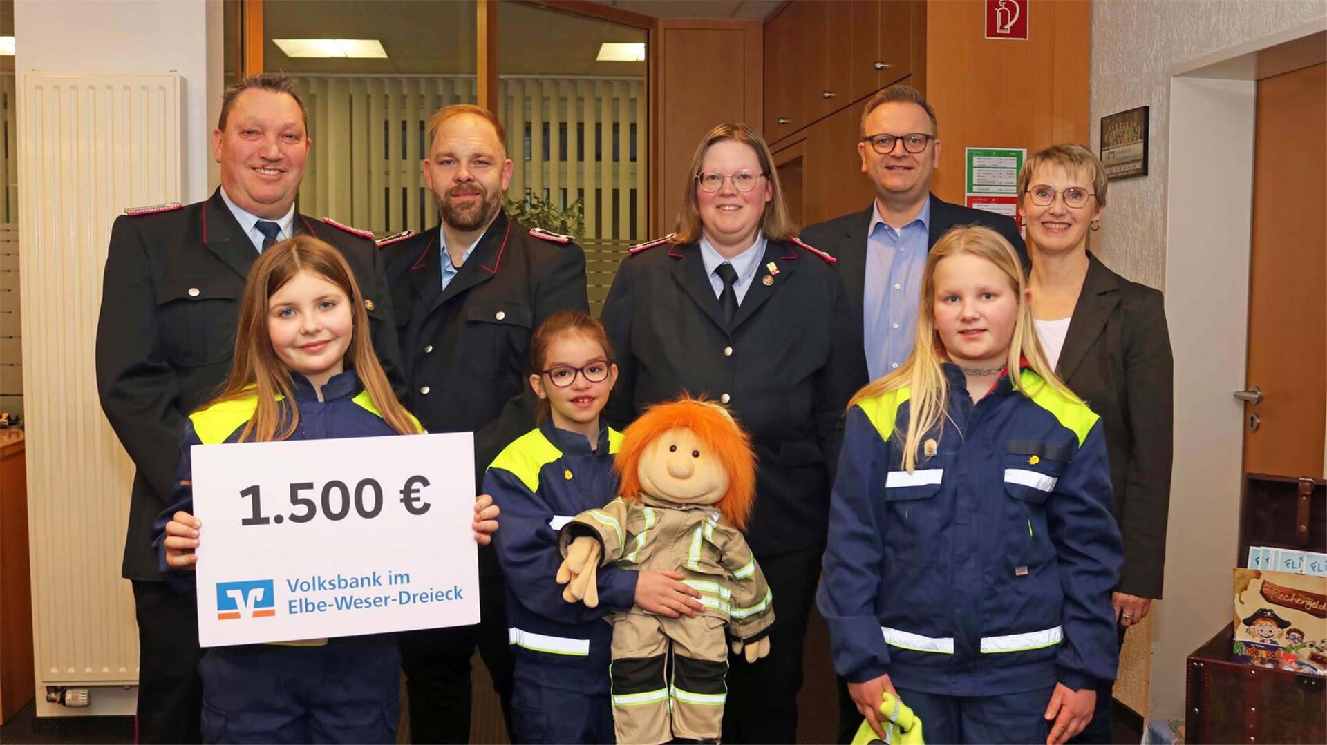  Drei Kinder in blauen und gelben Feuerwehruniformen stehen in der vorderen Reihe, eines hält ein Plakat mit der Aufschrift '1.500 € Volksbank im Elbe-Weser-Dreieck', ein anderes eine Stoffpuppe in Feuerwehrkleidung. Dahinter stehen fünf Erwachsene, darunter Männer in Feuerwehruniformen und zwei Personen in Zivilkleidung.