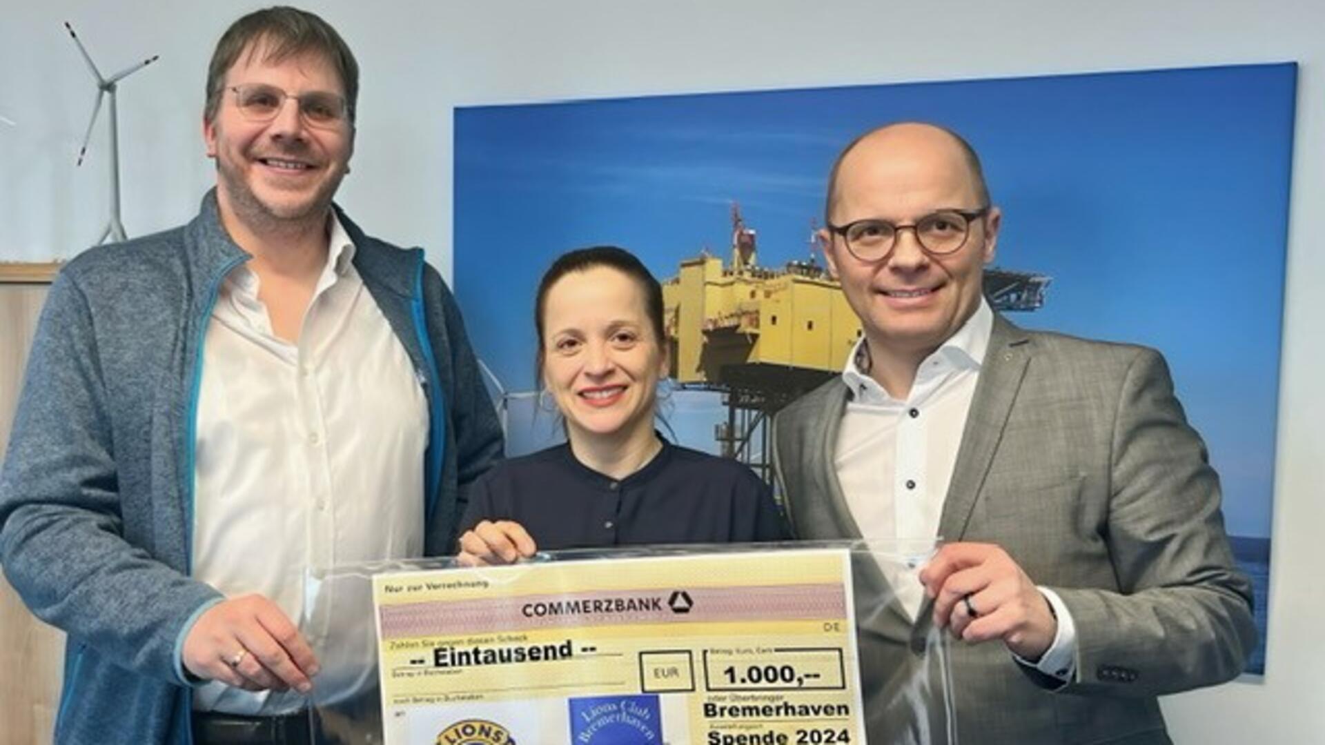 Drei Personen stehen nebeneinander und halten gemeinsam einen großen symbolischen Scheck über 1.000 Euro. Im Hintergrund ist ein Bild mit einer Offshore-Plattform und einer Windkraftanlage zu sehen.