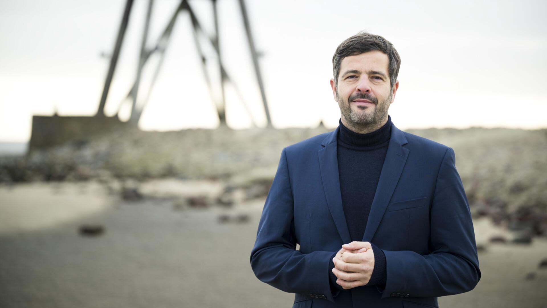 Porträtfoto von Daniel Schneider in Rollkragenpullover und Sakko am Strand