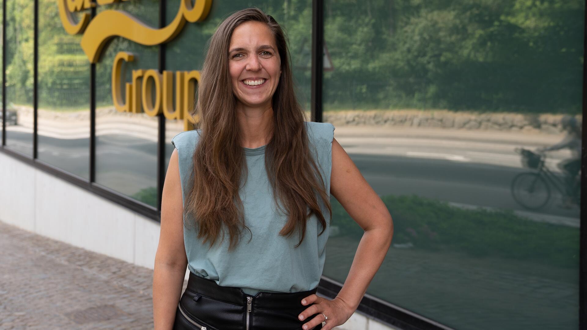 Eine Frau mit langen, dunklen Haaren steht vor einem Haus mit gläserner Fassade und lächelt in die Kamera.