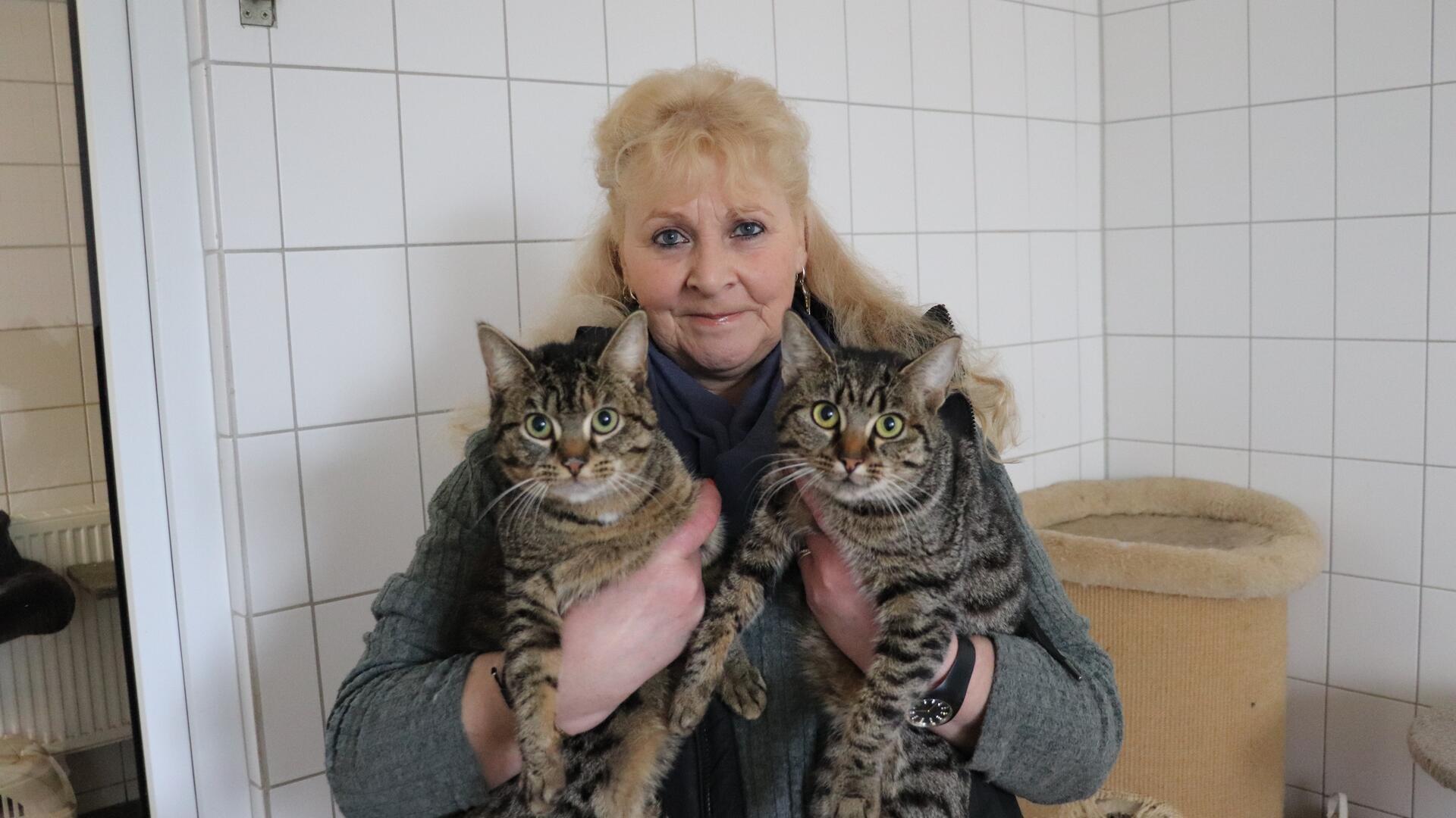 Eine Frau hält zwei Katzen im Arm und blickt in die Kamera.