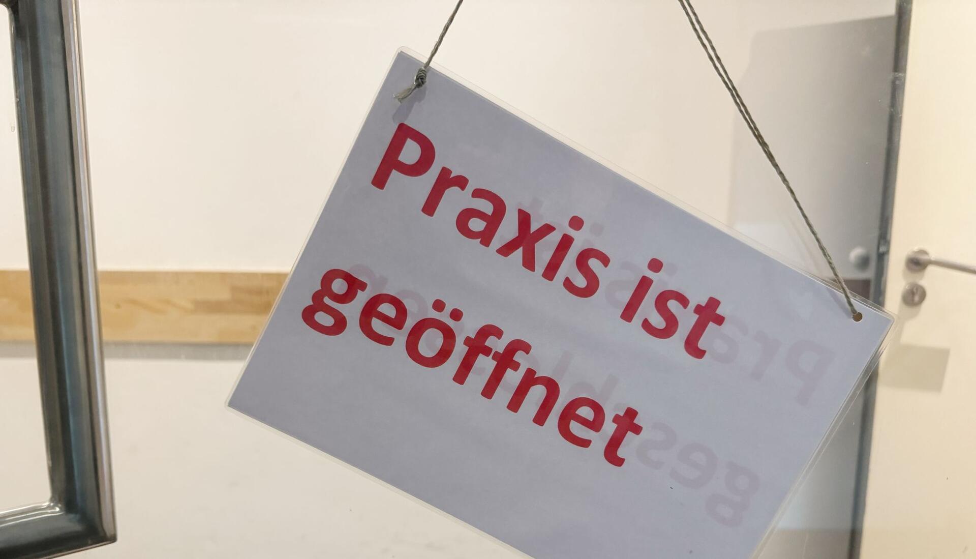 Ein Schild mit der Aufschrift „Praxis ist geöffnet" hängt an der Eingangstür einer Arztpraxis.