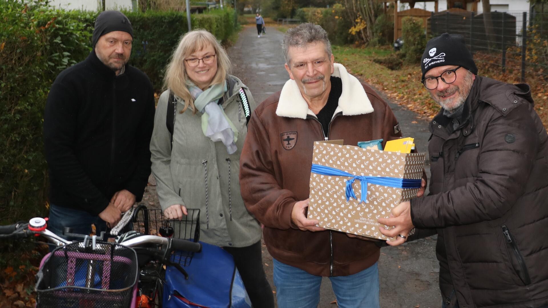 Drei Männer und eine Frau stehen auf einem Weg in herbstlicher Umgebung. Zwei von ihnen halten ein Fahrrad, während ein Mann dem anderen eine verpackte Geschenkbox mit blauer Schleife überreicht.