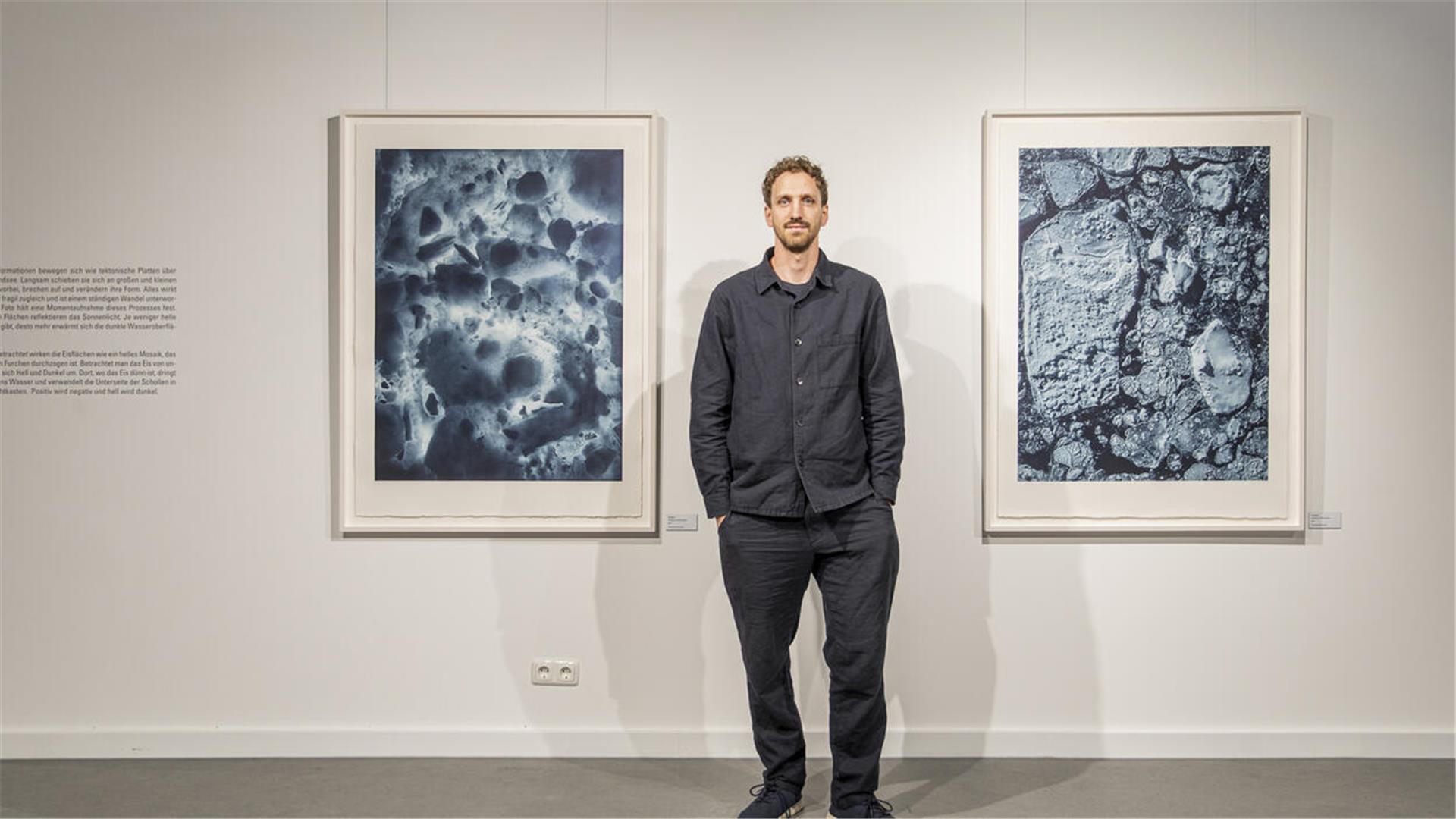 Ein Mann steht zwischen zwei großen Fotos, die viel blau und das Eismeer von unten (linkes Bild) und von oben zeigen.