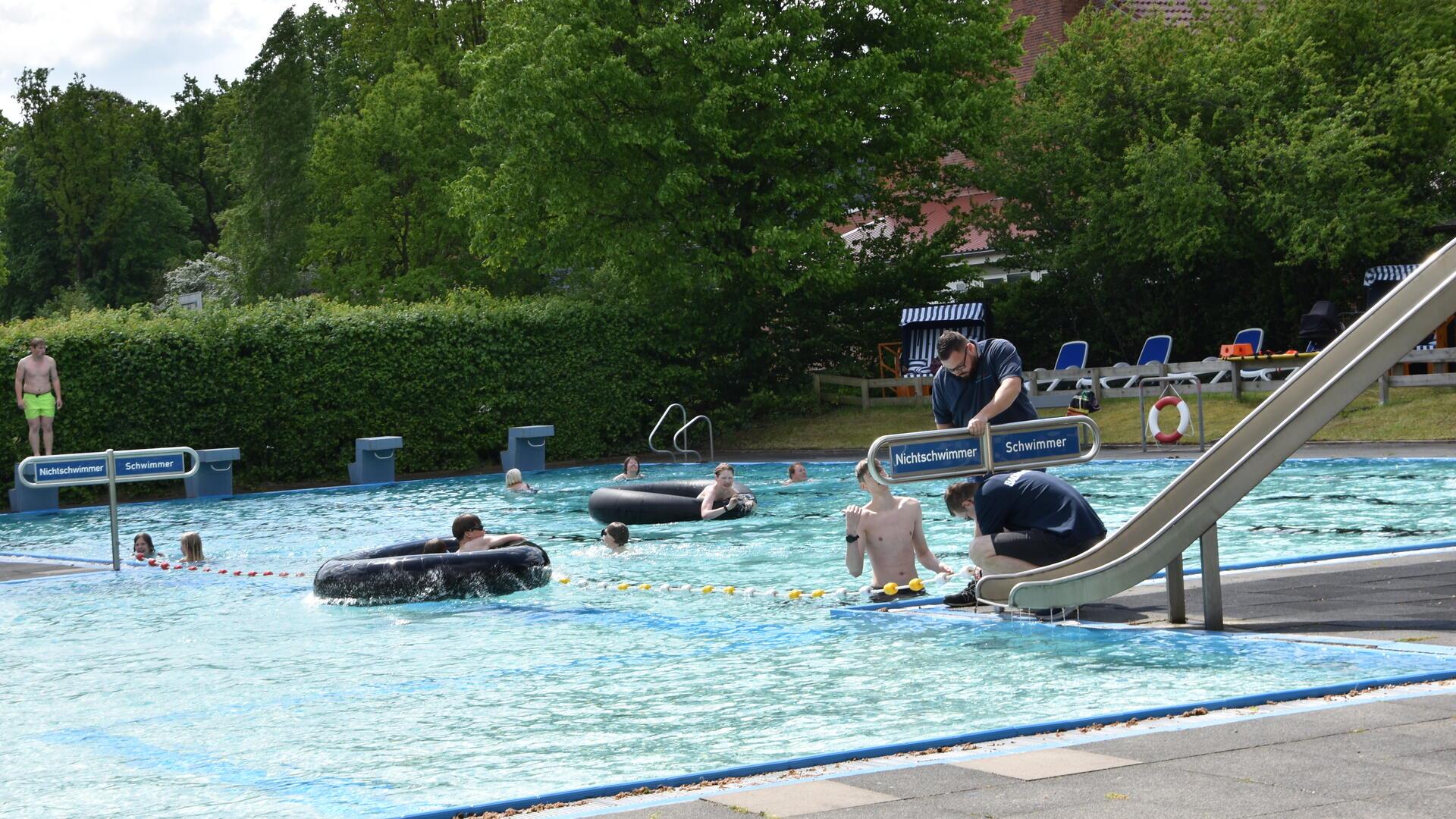 Ein Freibad mit mehreren Personen im Wasser. Einige Menschen benutzen große Schwimmringe, andere schwimmen oder stehen im flachen Wasser. Rechts im Bild befindet sich eine Wasserrutsche. Am unteren Ende montieren zwei Männer ein Schild, das das Becken in Nichtschwimmer und Schwimmer aufteilt. 