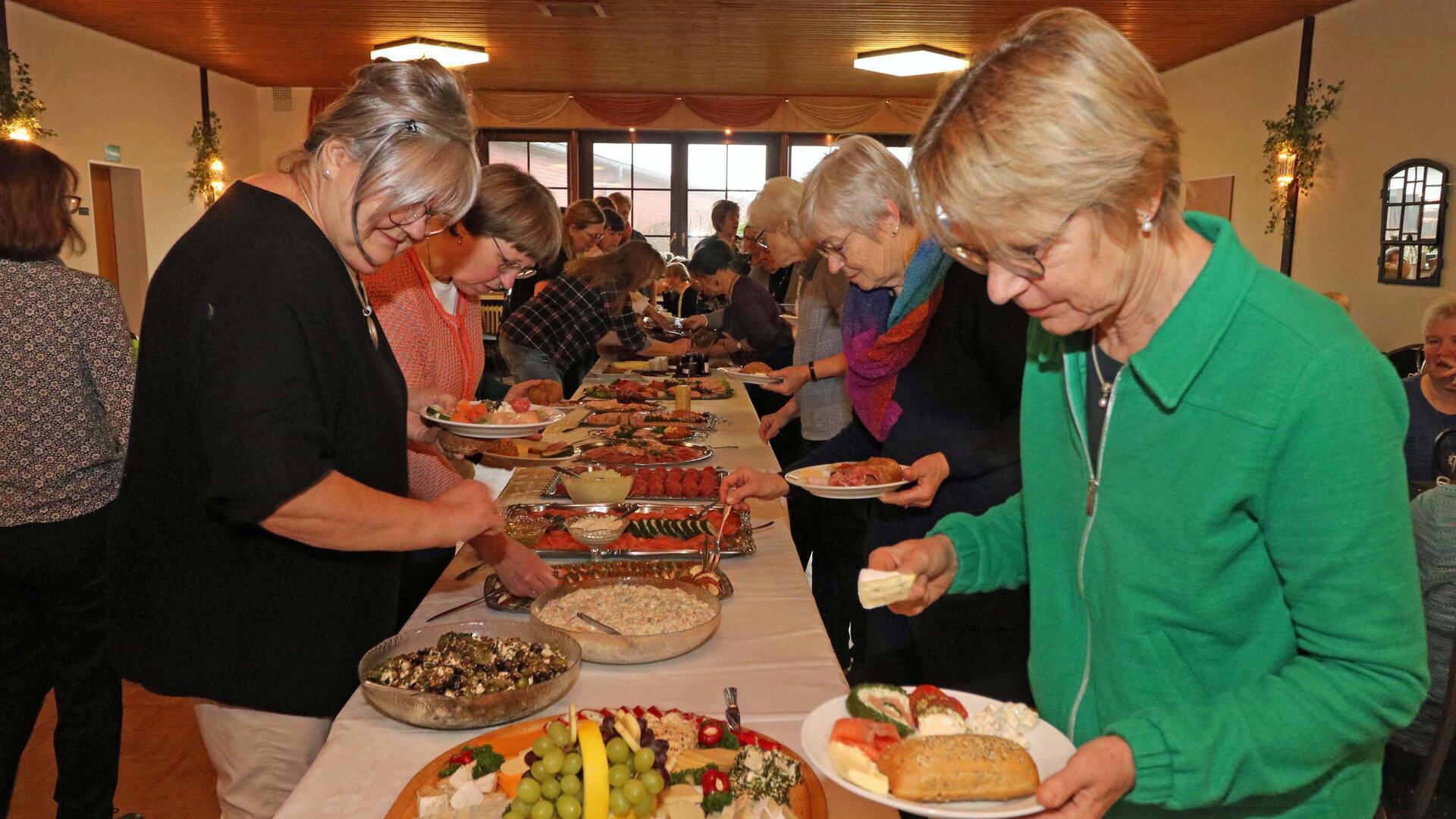 Eine Gruppe von Frauen steht an einem Buffet in einem Raum einer Gaststätte. Sie bedienen sich an einer großen Auswahl an Speisen, darunter Obst, Brot, Aufschnitt und verschiedene Salate, die auf einem langen Tisch präsentiert sind.